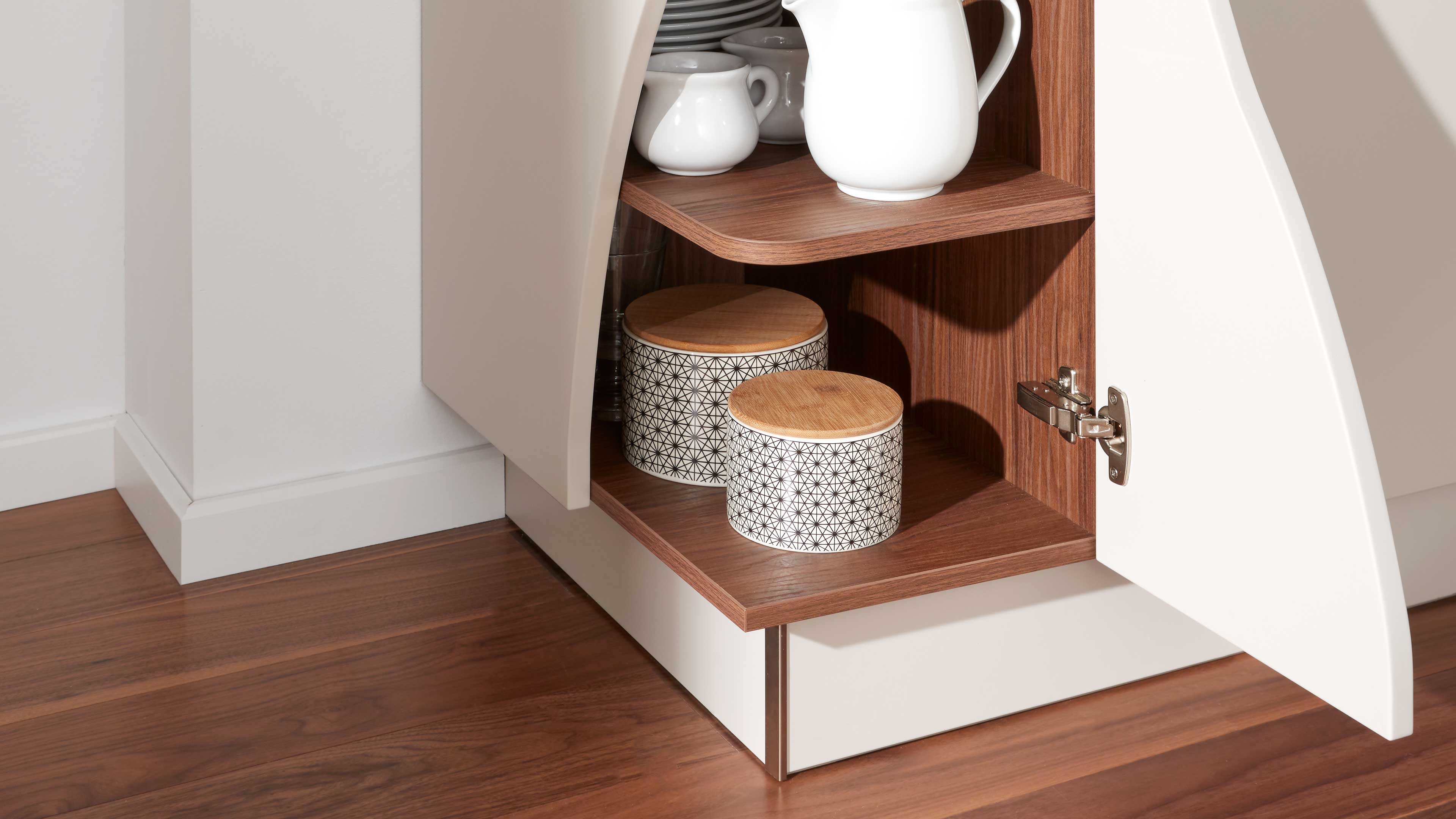 Geöffneter weißer Unterschrank mit warmholzfarbigen Innenflächen, zwei gemusterten Vorratsdosen mit Holzdeckeln auf dem unteren Brett.