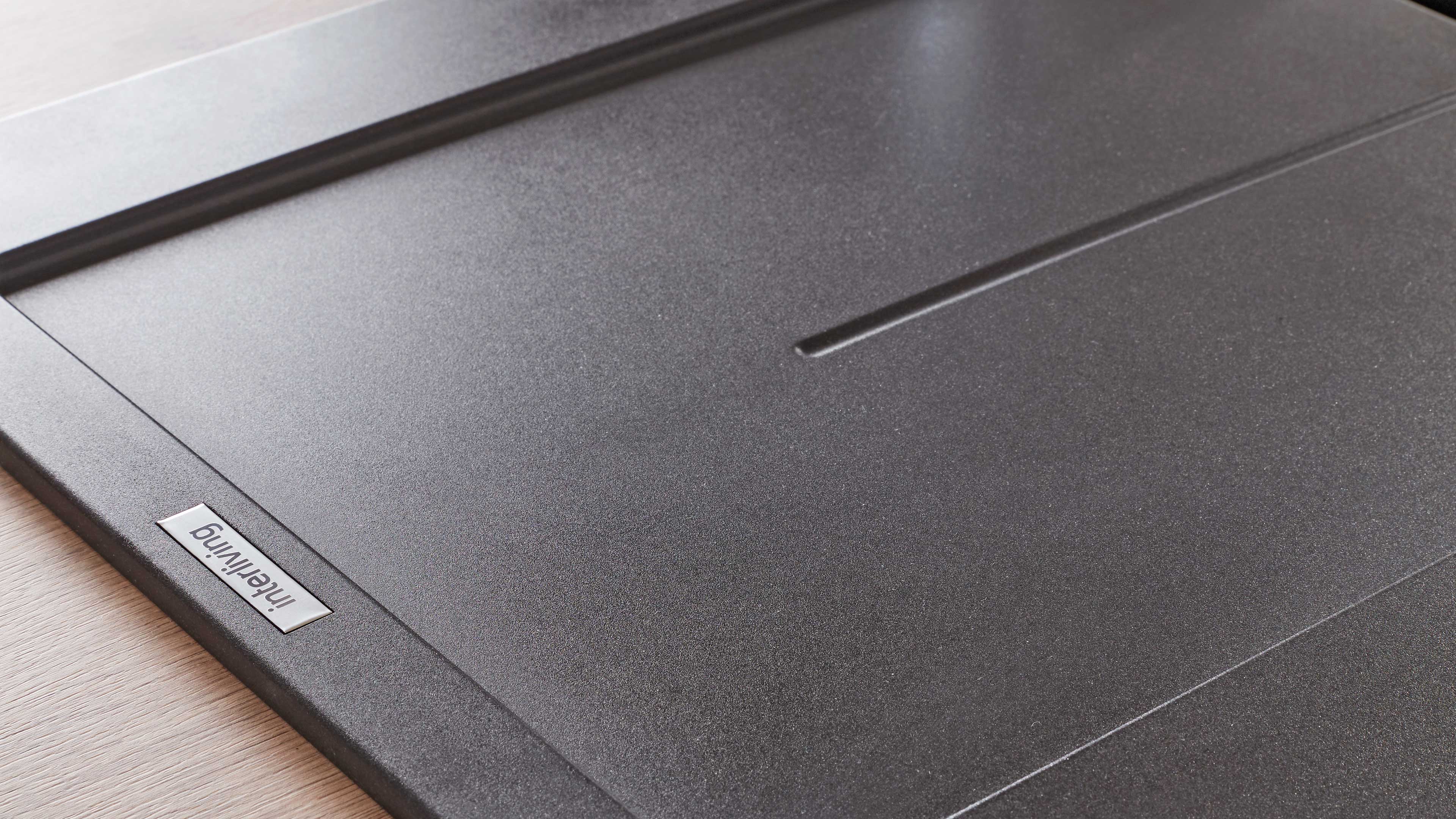 Nahaufnahme einer dunkelgrauen, fein strukturierten Möbeloberfläche mit eingeprägten Linien und kleinem Metallplättchen am Rand.