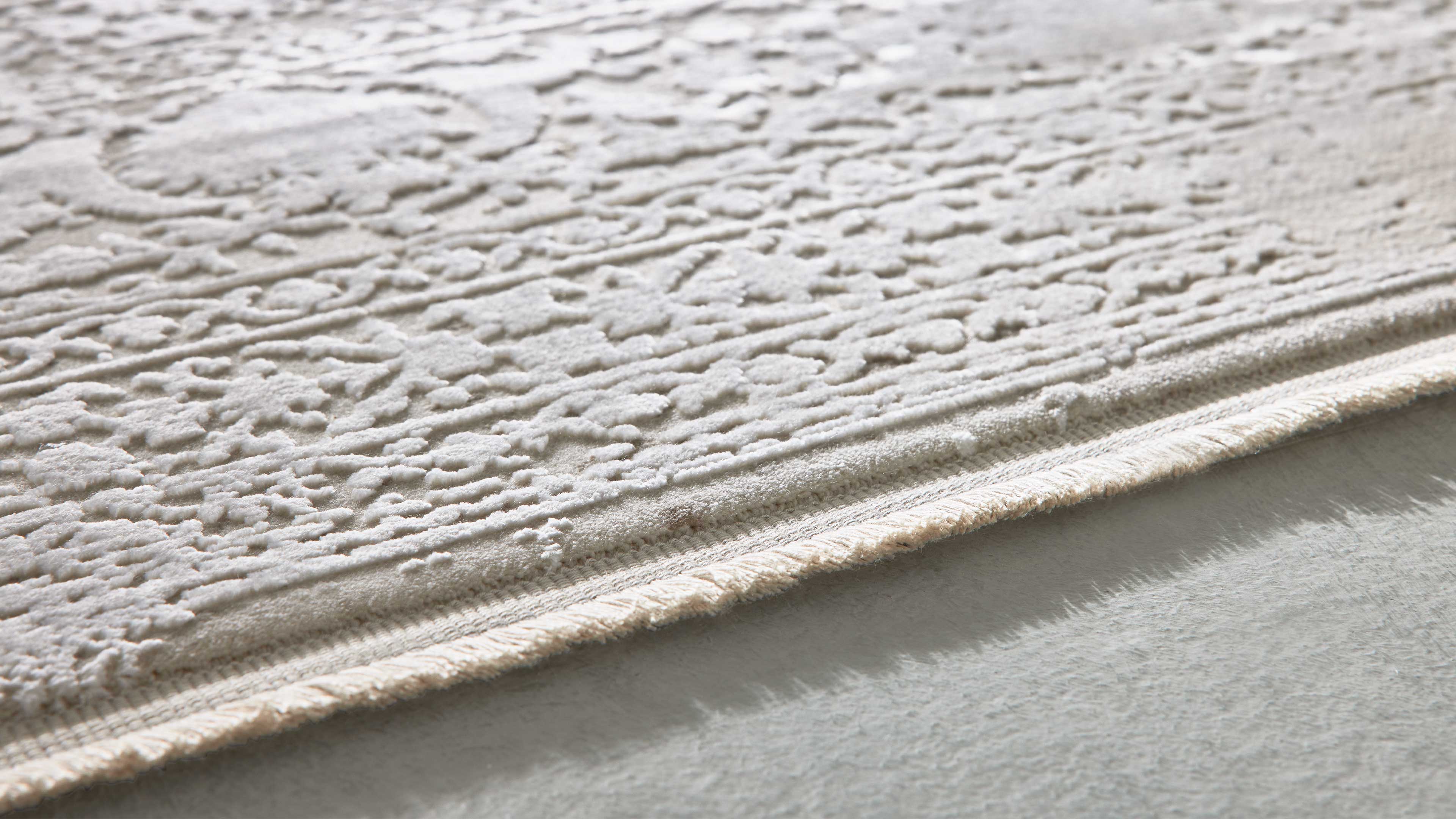 Nahaufnahme eines hellbeigen Teppichs mit erhabenen ornamentalen Mustern und sichtbarer umgenähter Kante auf hellem Boden.