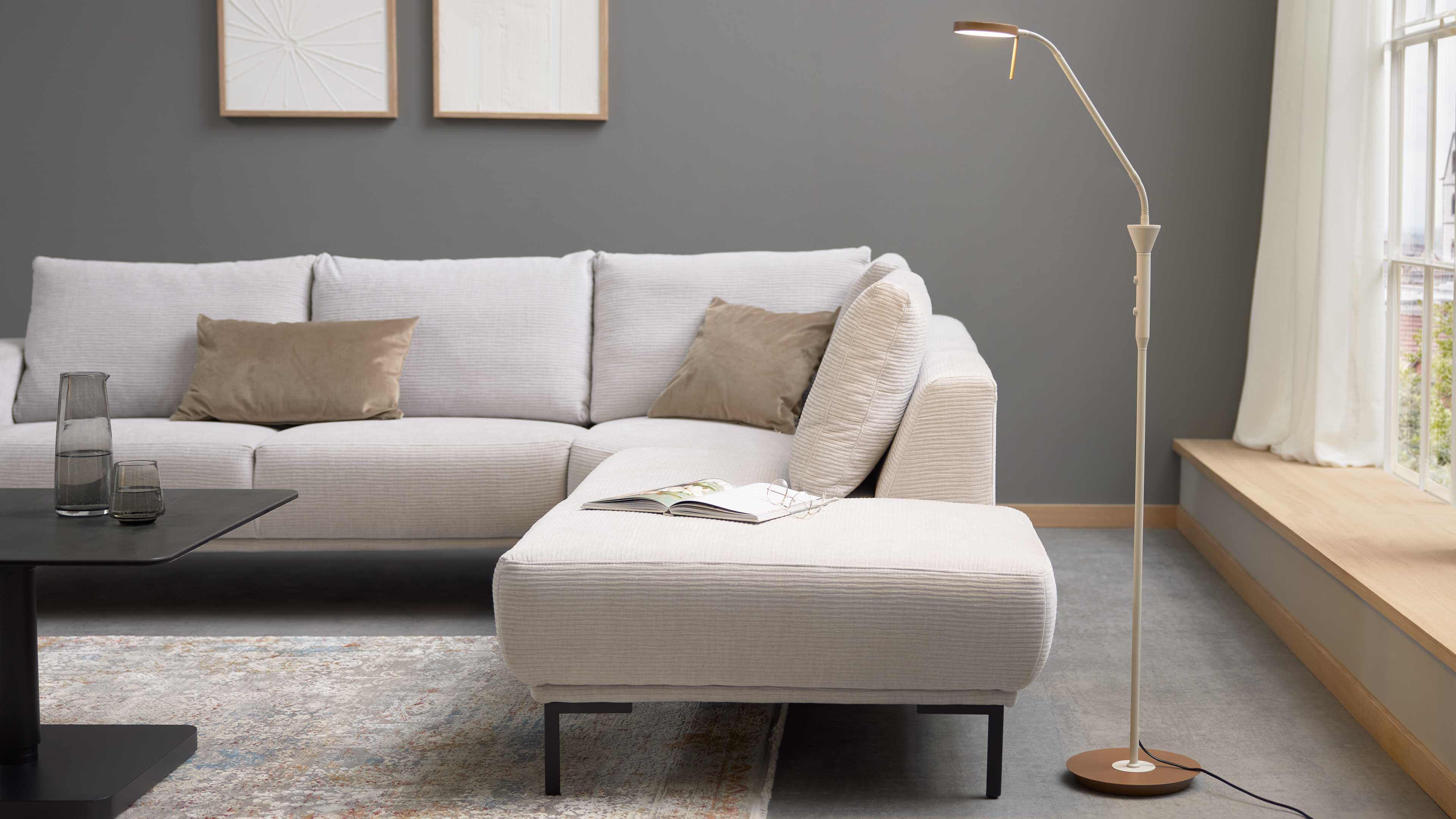Hellbeiges Ecksofa mit Ottomane, zwei beige Kissen und offenes Magazin mit Brille auf dem Sitz; rechts schlanke Stehlampe auf rundem Sockel.
