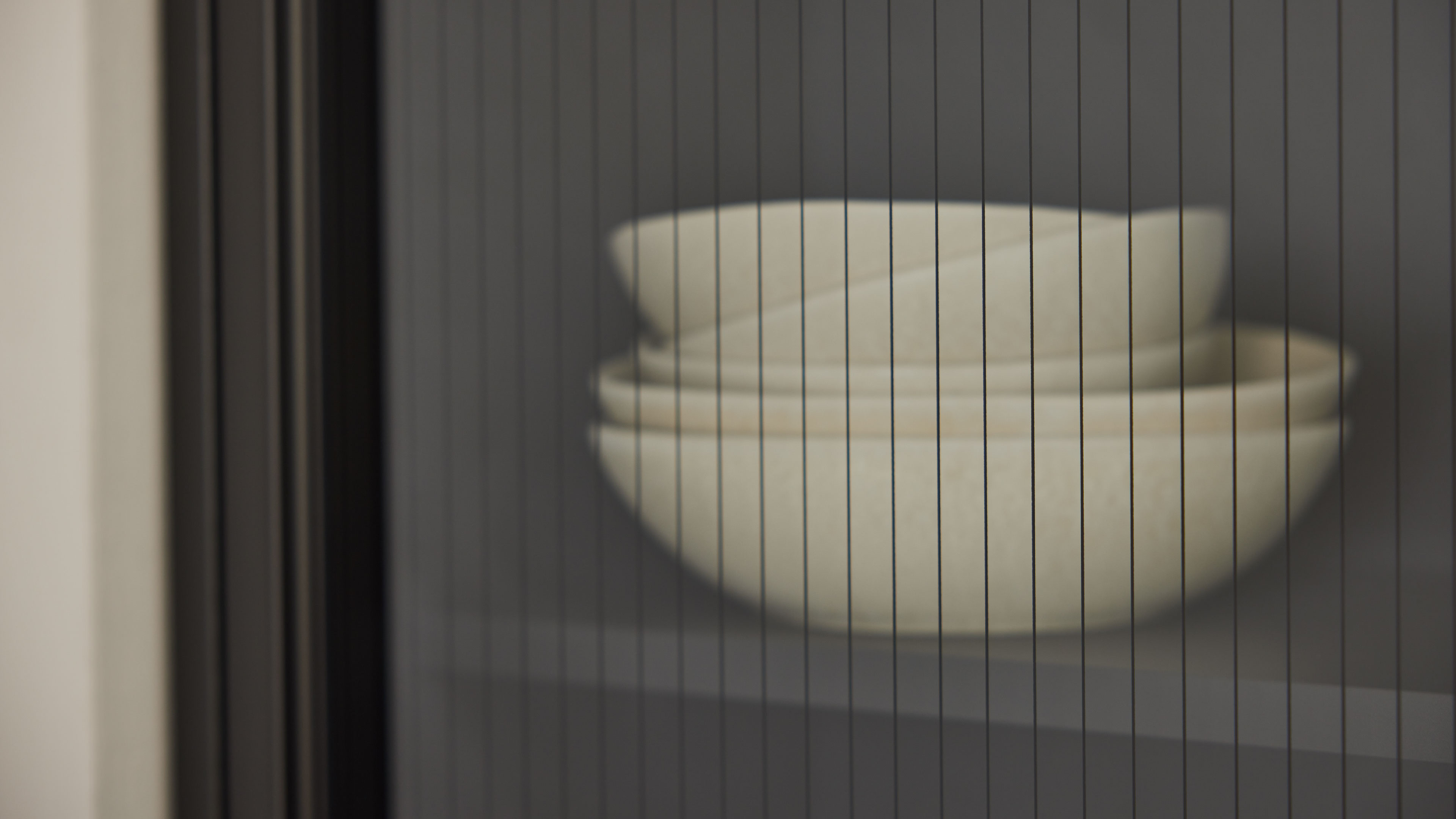 Gestapelte weiße Schalen auf einem Regalbrett, durch eine mit feinen vertikalen Linien versehene Glasscheibe und dunklen Rahmen sichtbar.