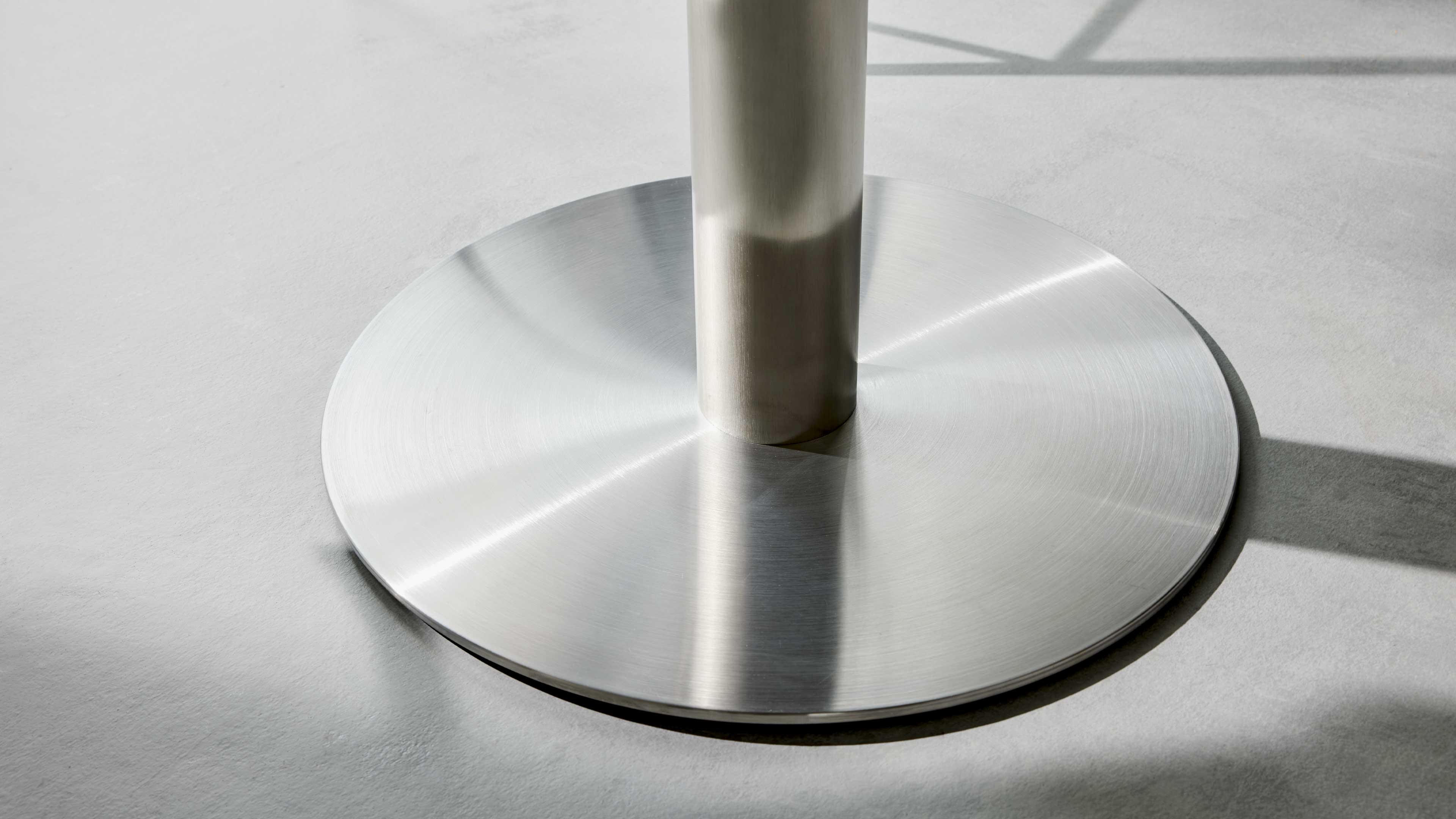 Rundes, gebürstetes Metallfußteil mit zylindrischer Stütze steht auf hellem Betonboden, sichtbare Schatten und Lichtreflexe.