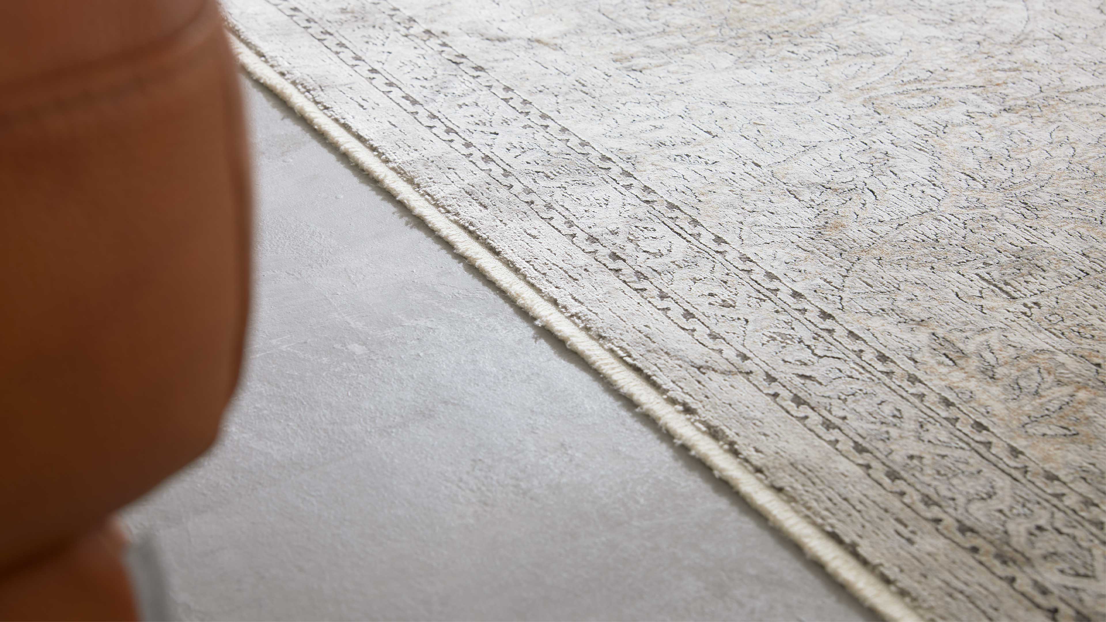 Heller beige Teppich mit feiner ornamentaler Bordüre liegt auf glattem hellgrauen Boden, links unscharf ein braunes Polsterelement.