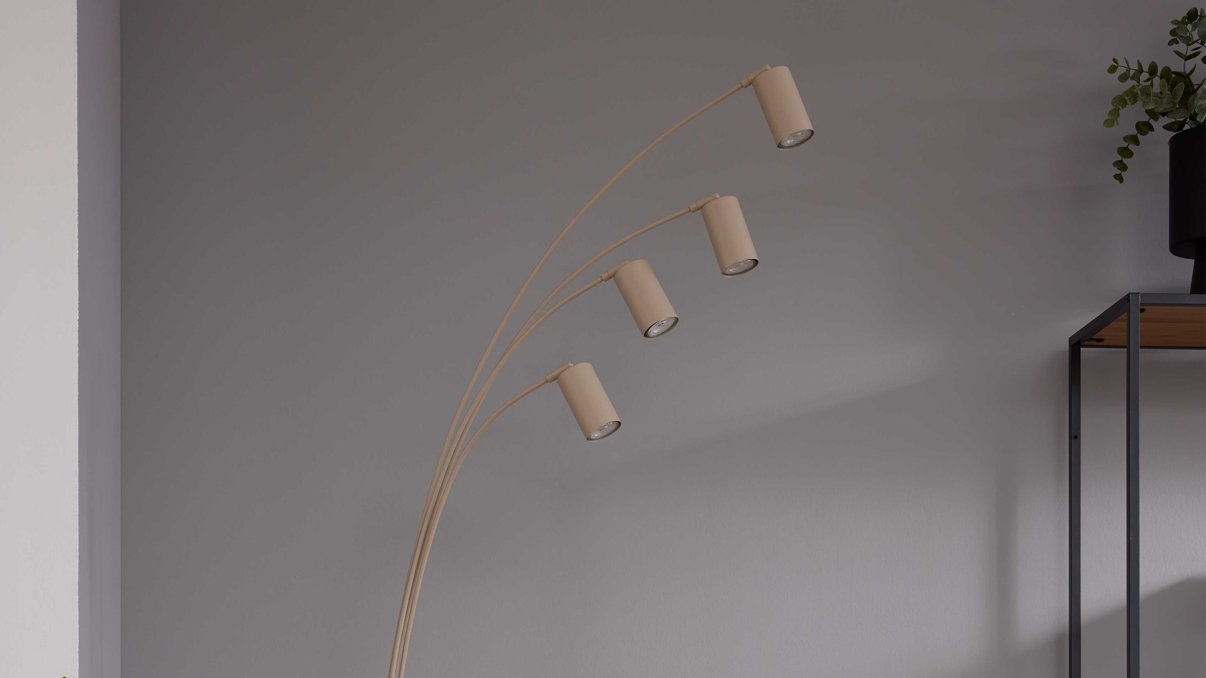 Stehende Bogenlampe mit vier beigen, zylindrischen Leuchtenarmen vor einer grauen Wand, rechts schmales Metallregal mit Pflanze.