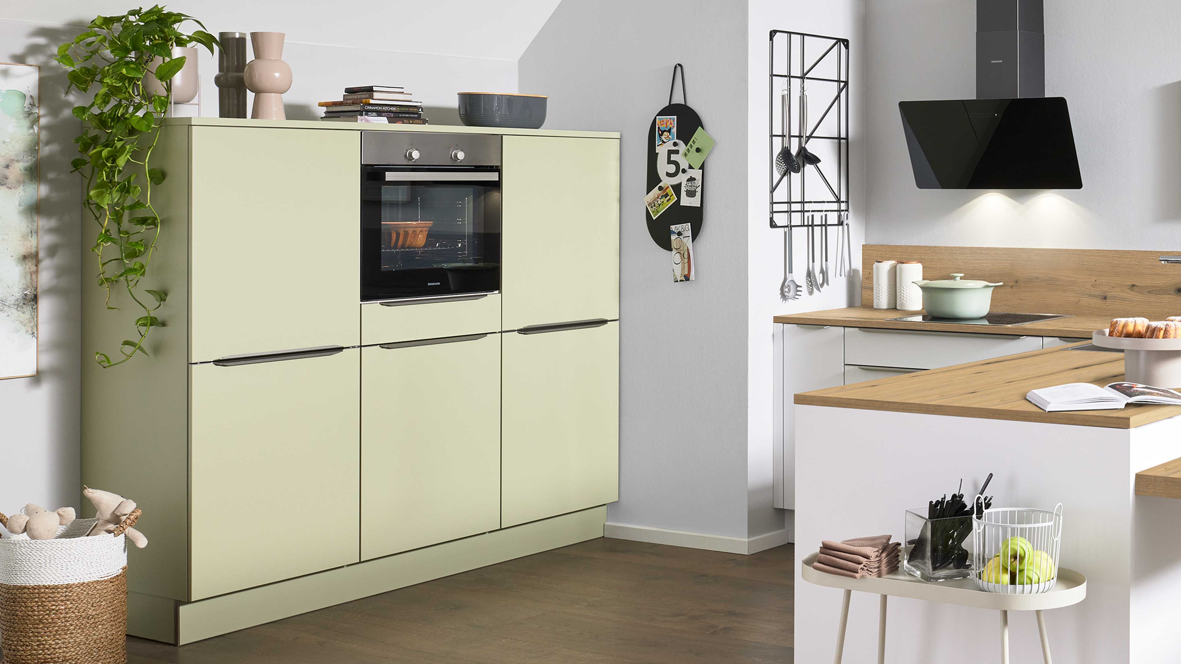 Hellgrüne Hochschrankwand mit eingebautem Backofen und Pflanze oben; rechts Holzarbeitsplatte mit Dunstabzug und hängenden Utensilien.