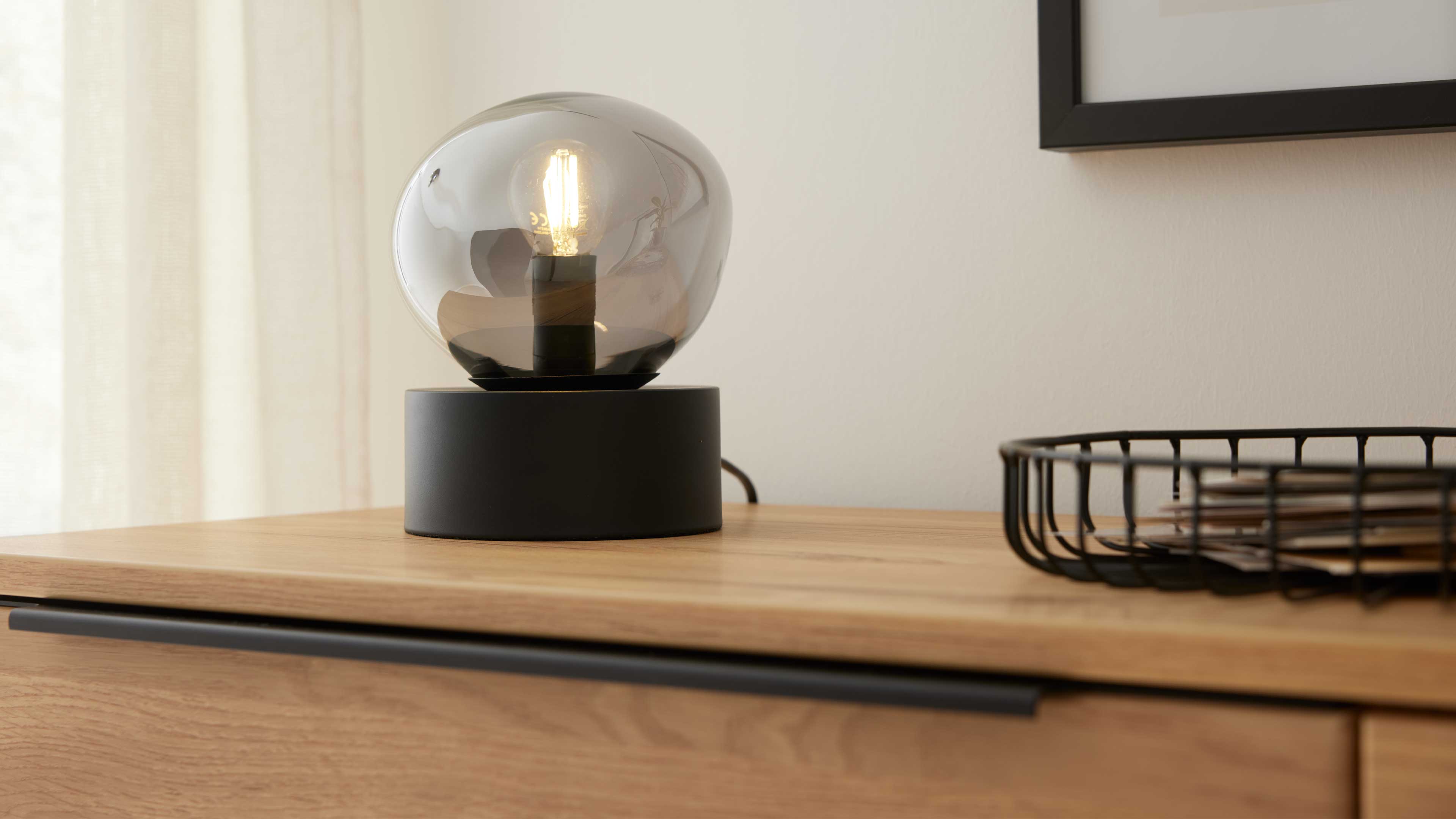 Auf einer Holzkommode steht eine schwarze Tischlampe mit rundem rauchglas-Schirm und sichtbarer Glühbirne, rechts ein Drahtkorb mit Papieren.