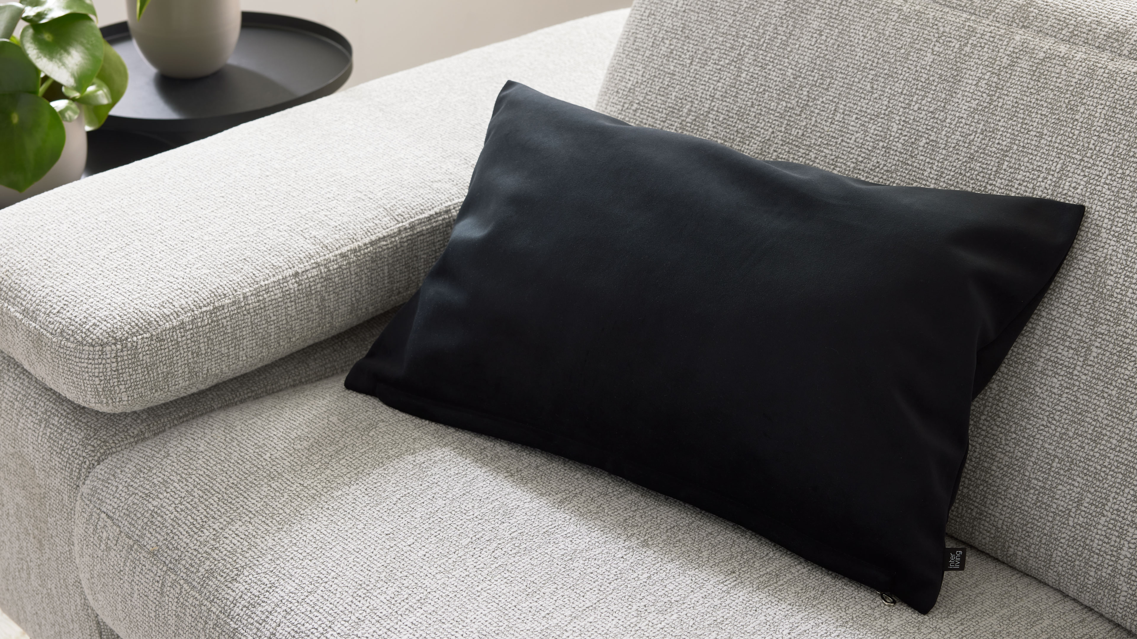 Helles, grob gewebtes Sofa mit schwarzem rechteckigem Kissen auf Sitzfläche, links angeschnittener Beistelltisch mit Pflanze.