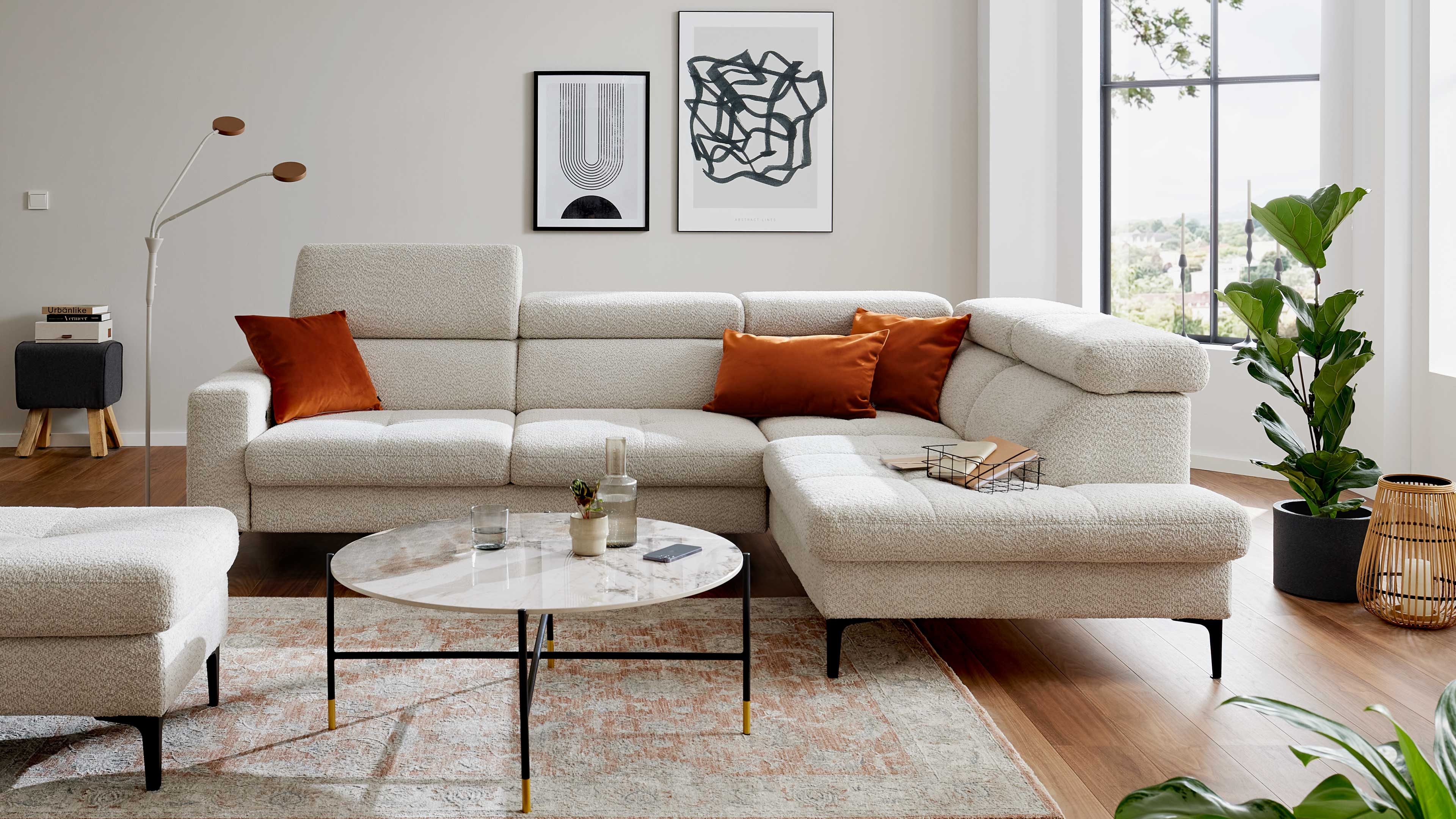 Großes hellbeiges Ecksofa mit drei rostfarbenen Kissen vor rundem Marmortisch, Wandbildern, Stehlampe und Zimmerpflanze.