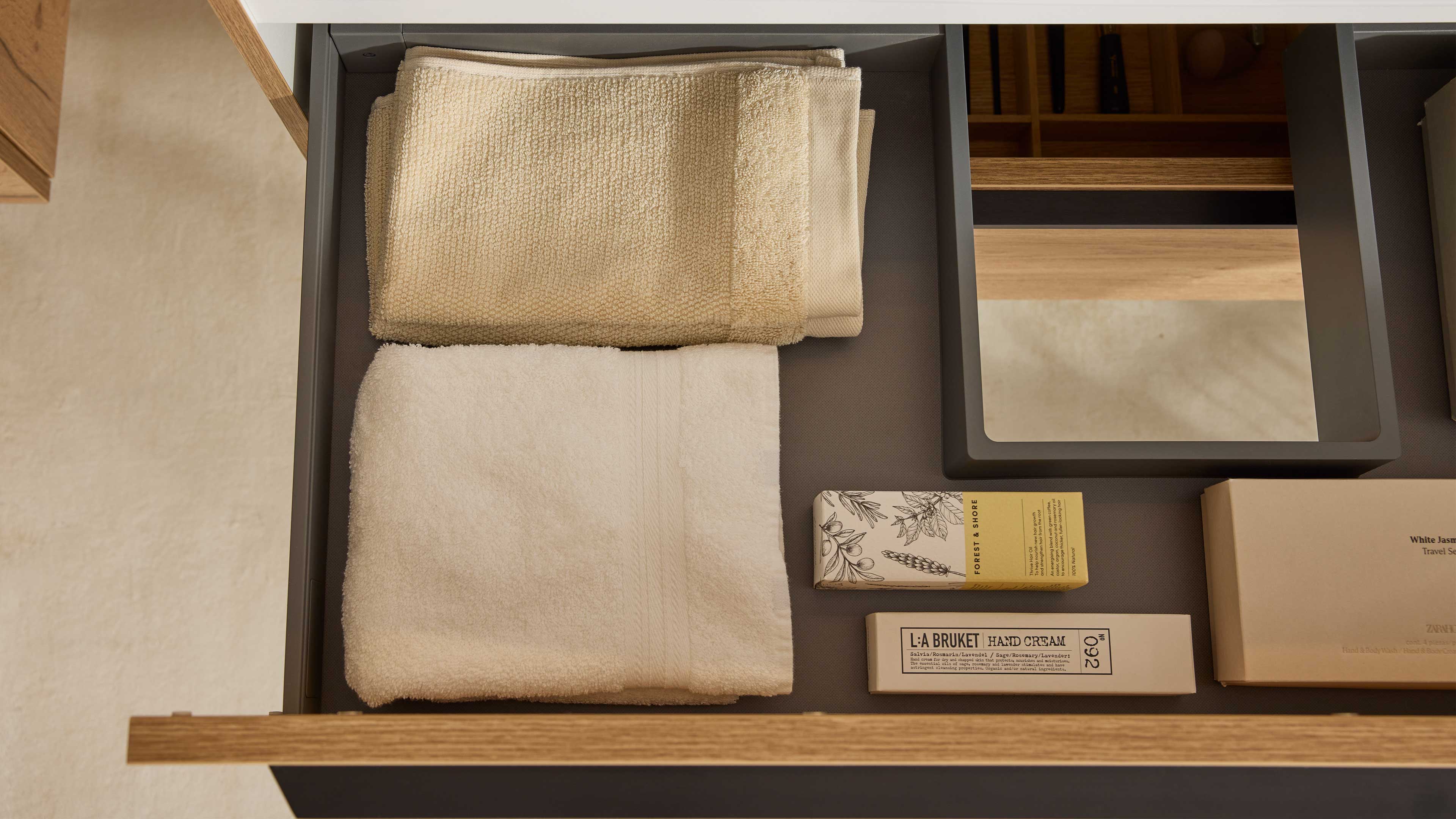 Offene Holzschublade mit grauem Innenfach, zwei gefalteten Handtüchern links und rechts kleine Kosmetikschachteln.