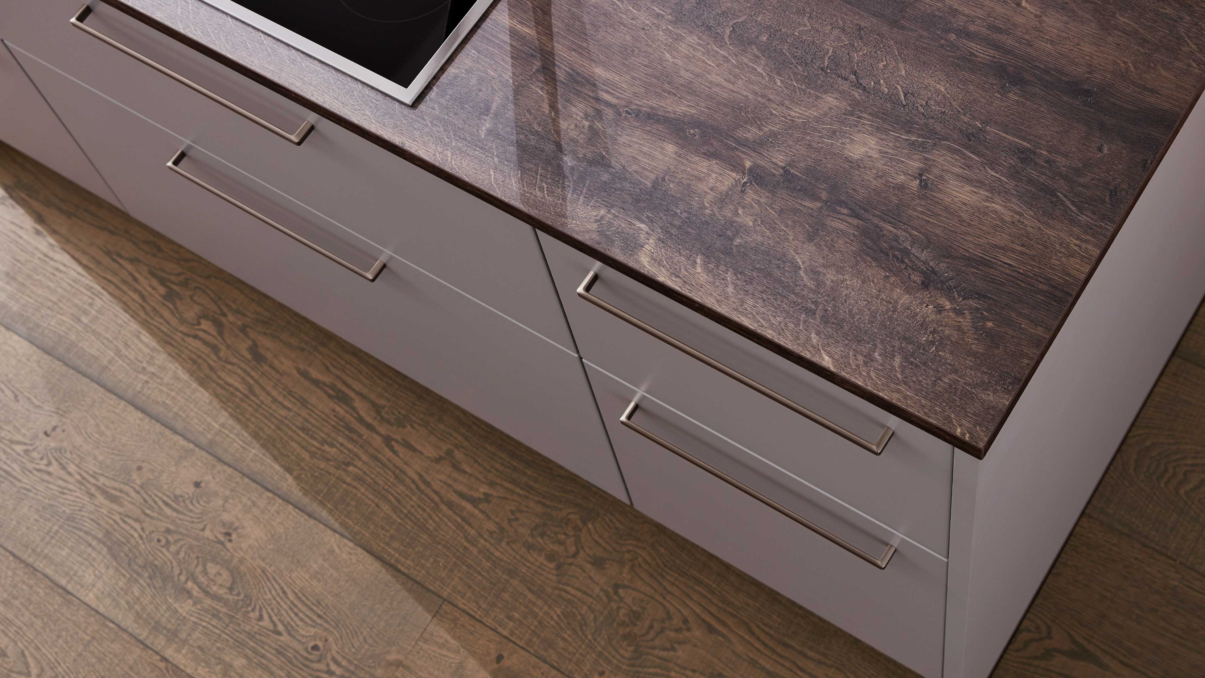 Graue Küchenschubladen mit langen Metallgriffen unter einer dunkel gezeichneten Holzarbeitsplatte, Teil eines Kochfelds sichtbar.