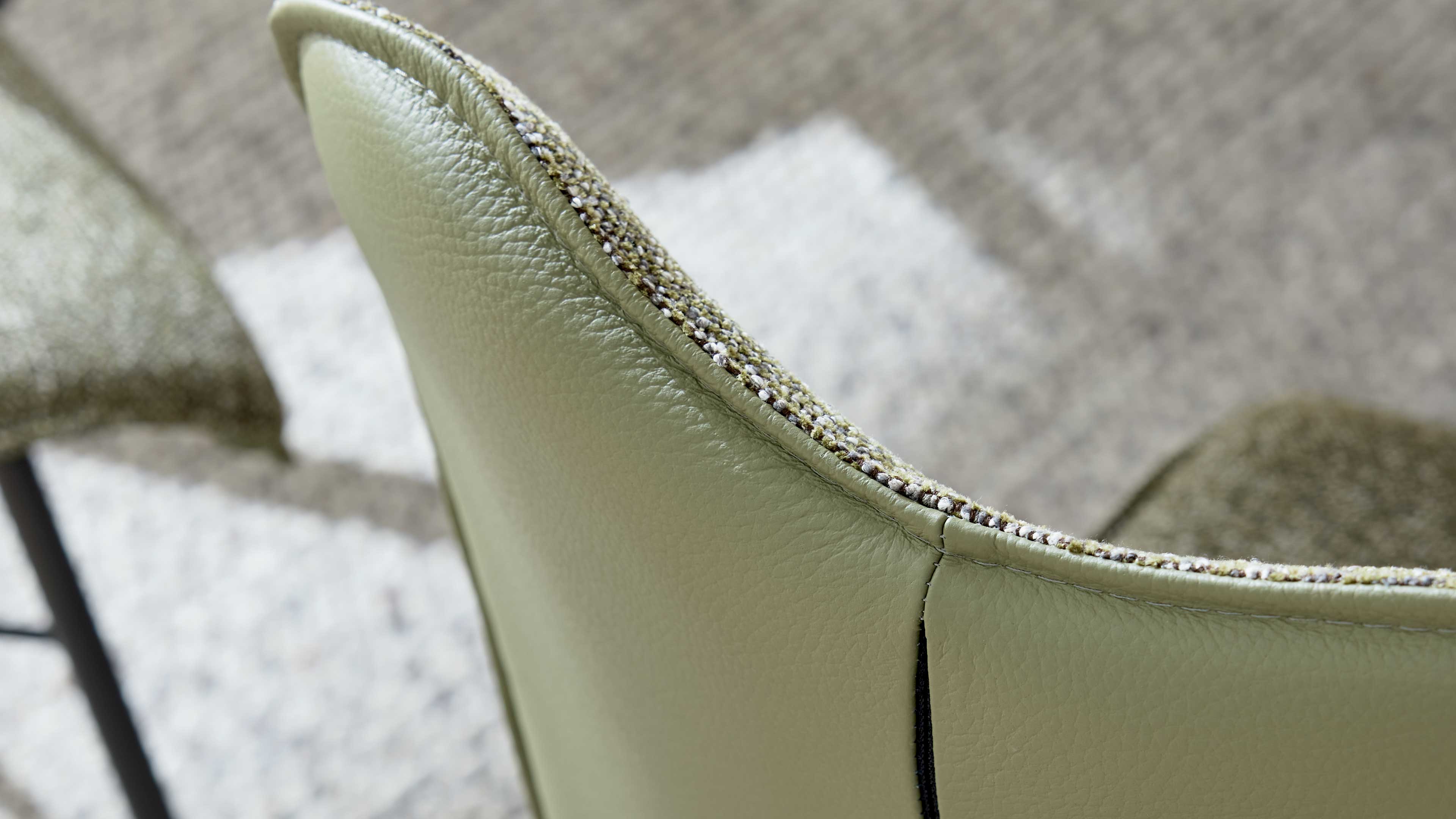 Rückenlehne eines hellgrünen Lederstuhls mit sichtbarer Naht und kontrastierendem Stoffbesatz, unscharfer Teppich im Hintergrund.