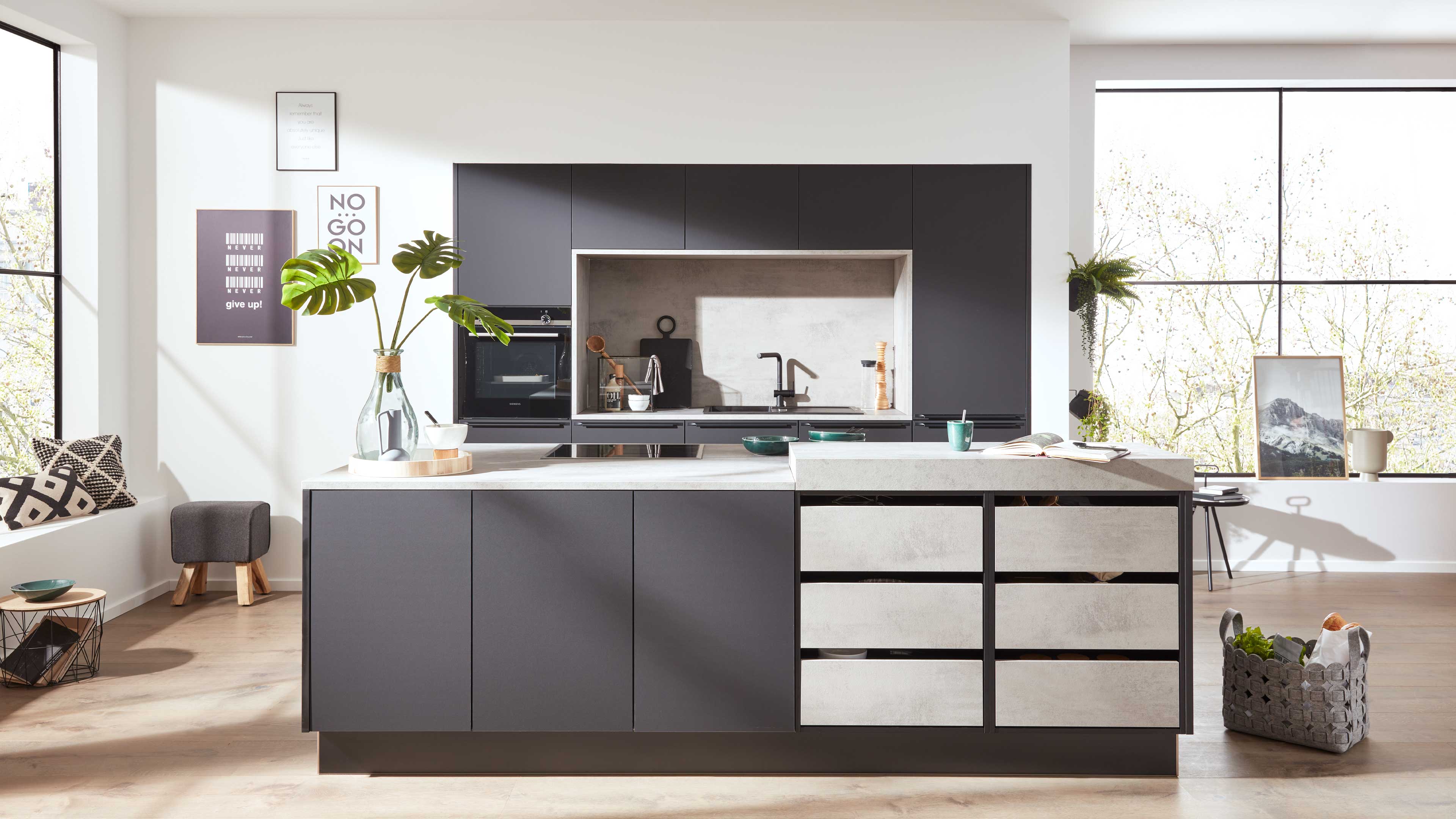 Moderne Küche mit großer dunkelgrauer Insel und Beton-Optik-Arbeitsplatte, Monstera-Vase, eingelassenem Spülbereich und großen Fenstern.