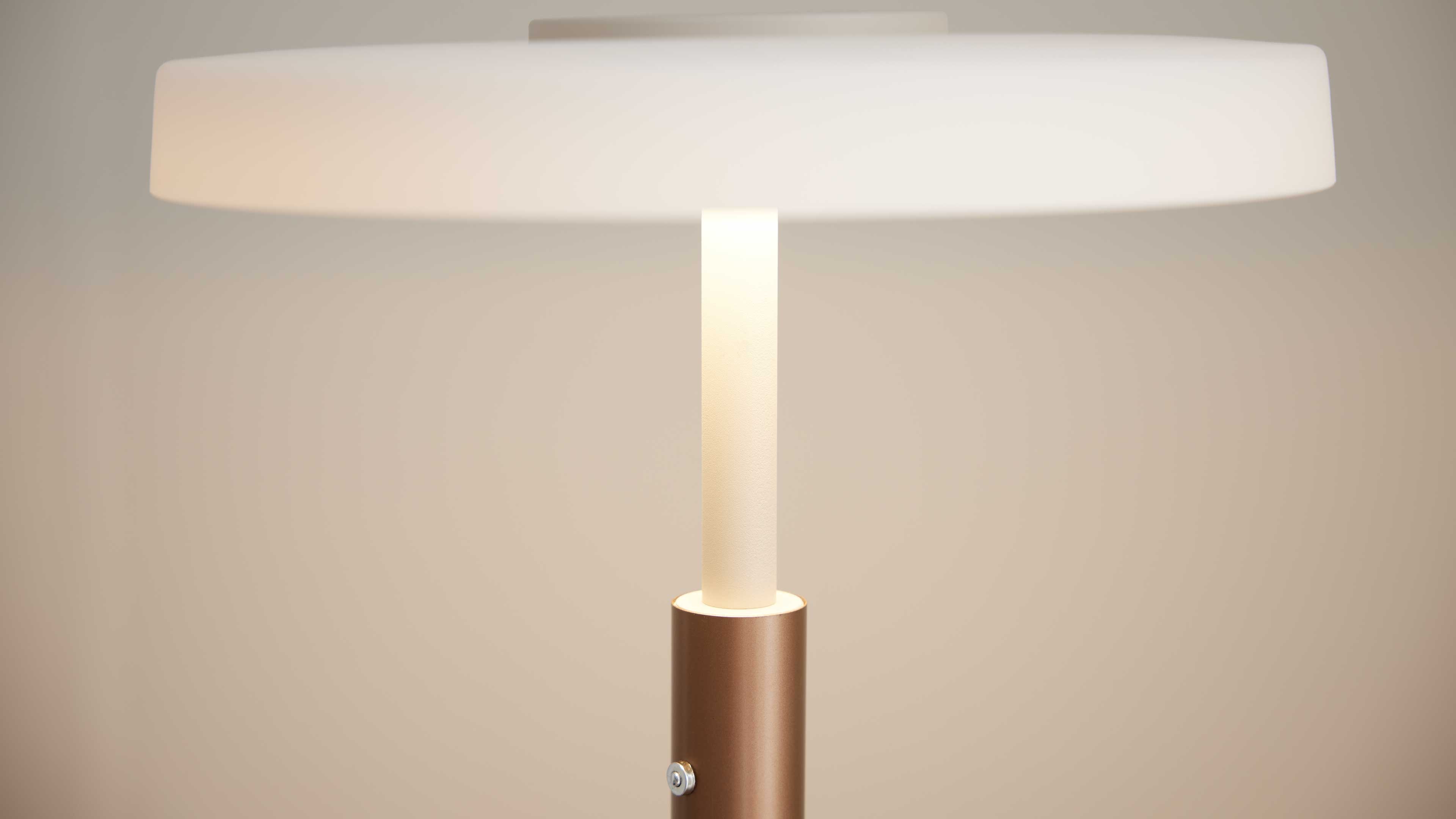 Tischlampe mit flachem weißen Schirm, zentralem opal-weißem Leuchtstab und bronzefarbenem zylindrischem Fuß mit sichtbarer Schraube.