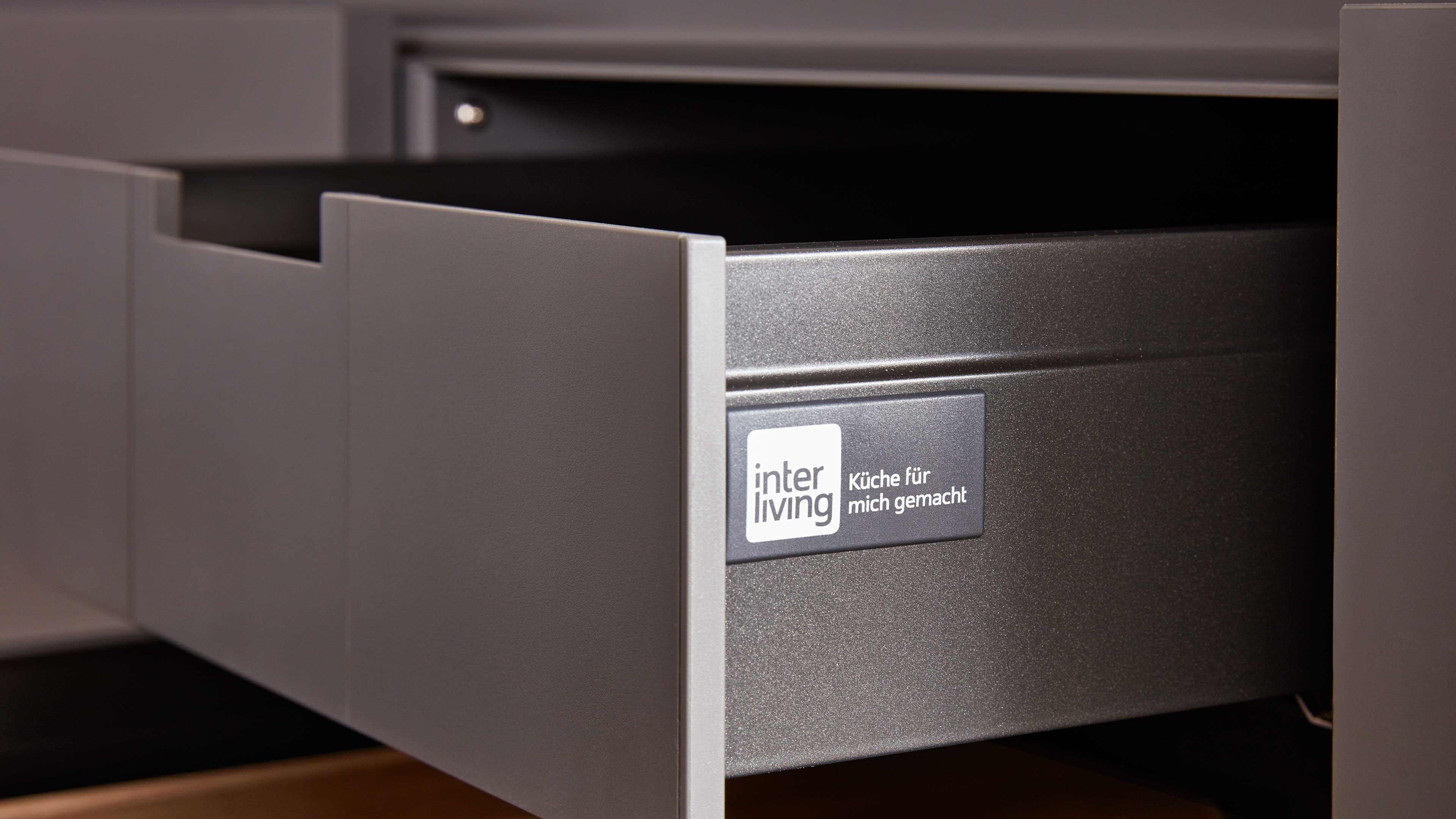 Offene graue Küchenschublade mit Griffausschnitt, metallischem Innenkasten und aufgesetztem Etikett 'interliving – Küche für mich gemacht'.