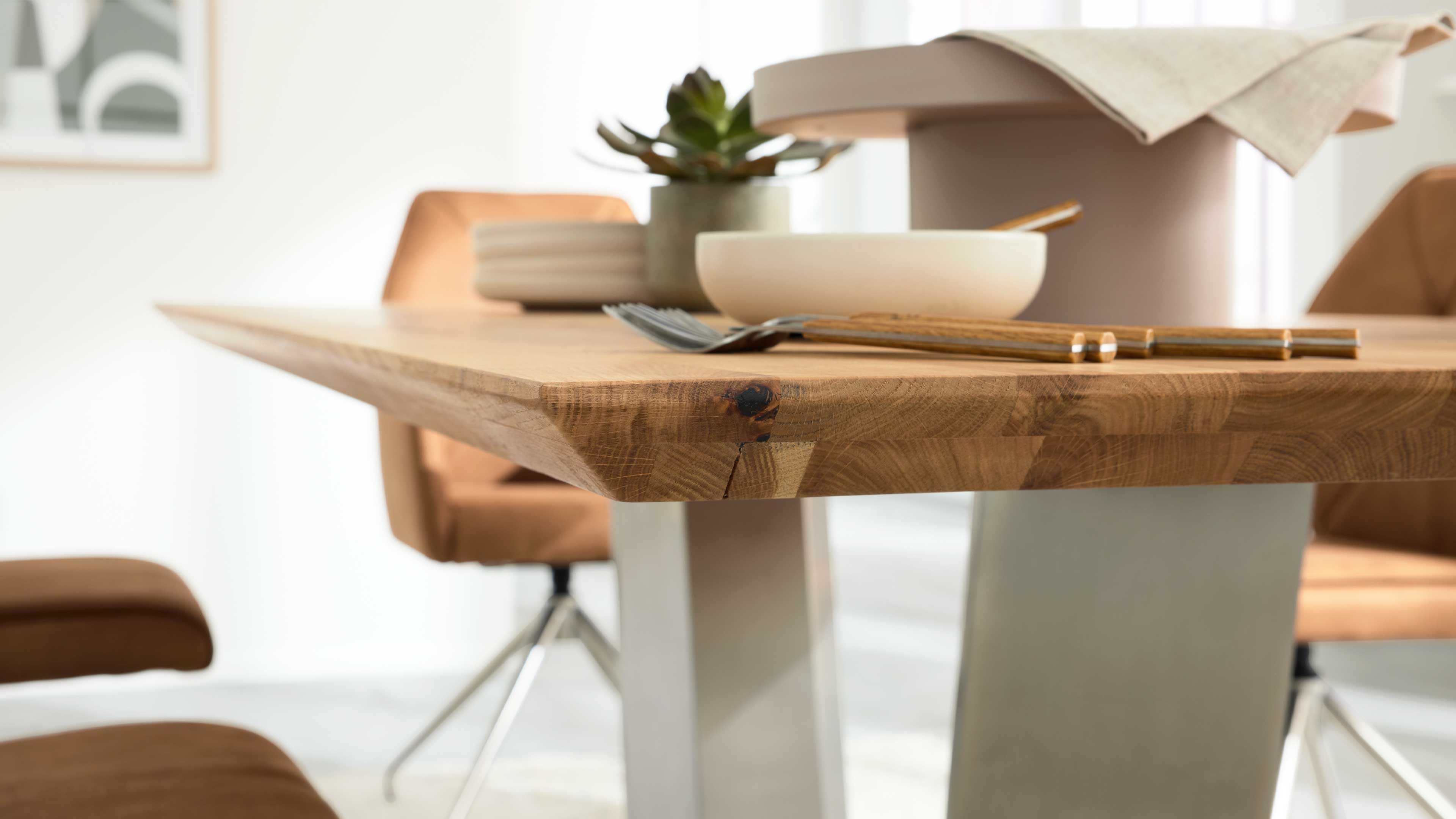 Holz-Tischkante mit sichtbarer Maserung, darauf Schüssel, Besteck und kleiner Sukkulenten-Topf; im Hintergrund Stuhl und Metallbein.