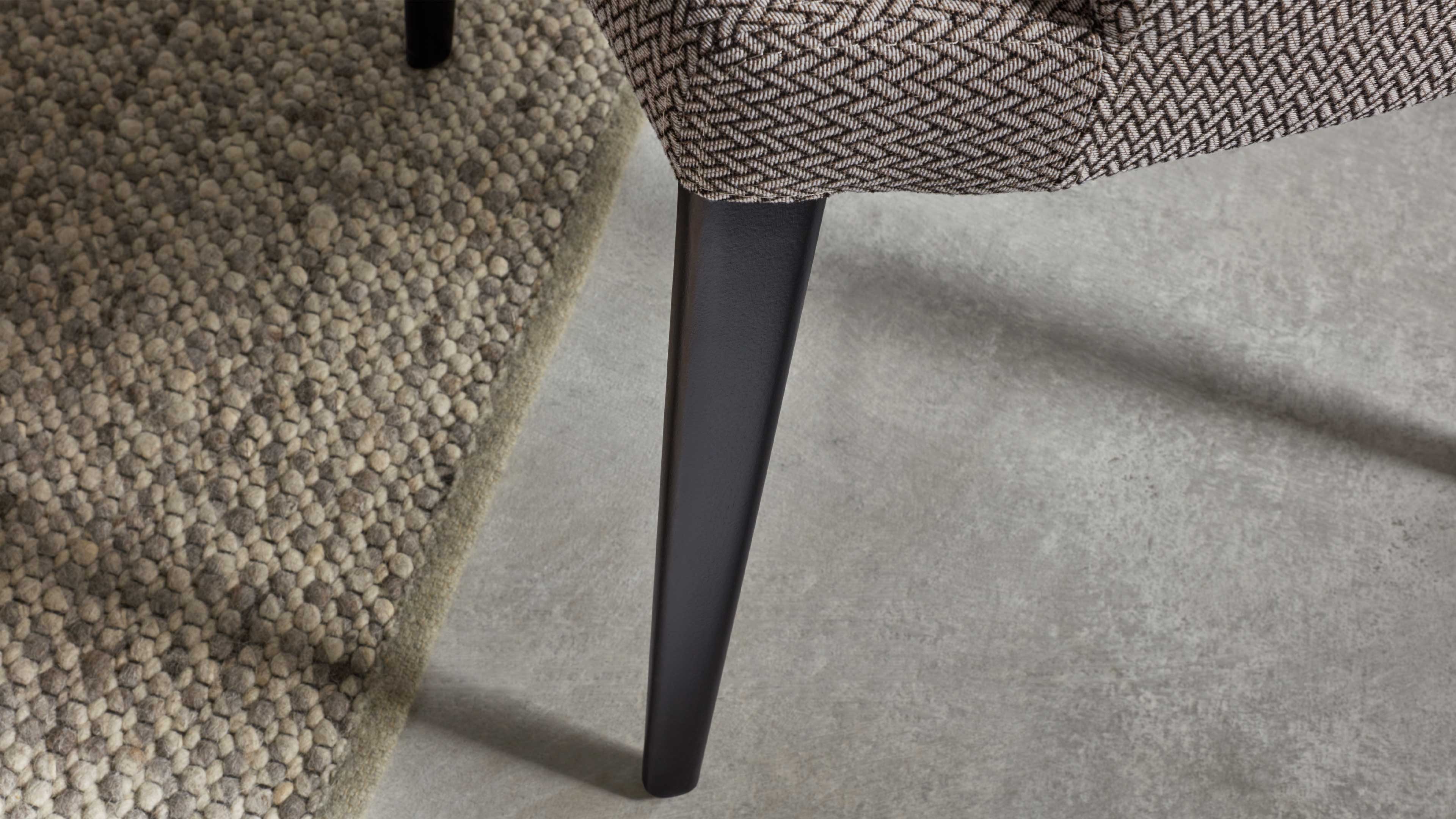 Schwarzes, konisch zulaufendes Möbelbein an beiger, zweifarbiger Polsterkante; links sichtbarer körniger Teppich, rechts grauer Betonboden.