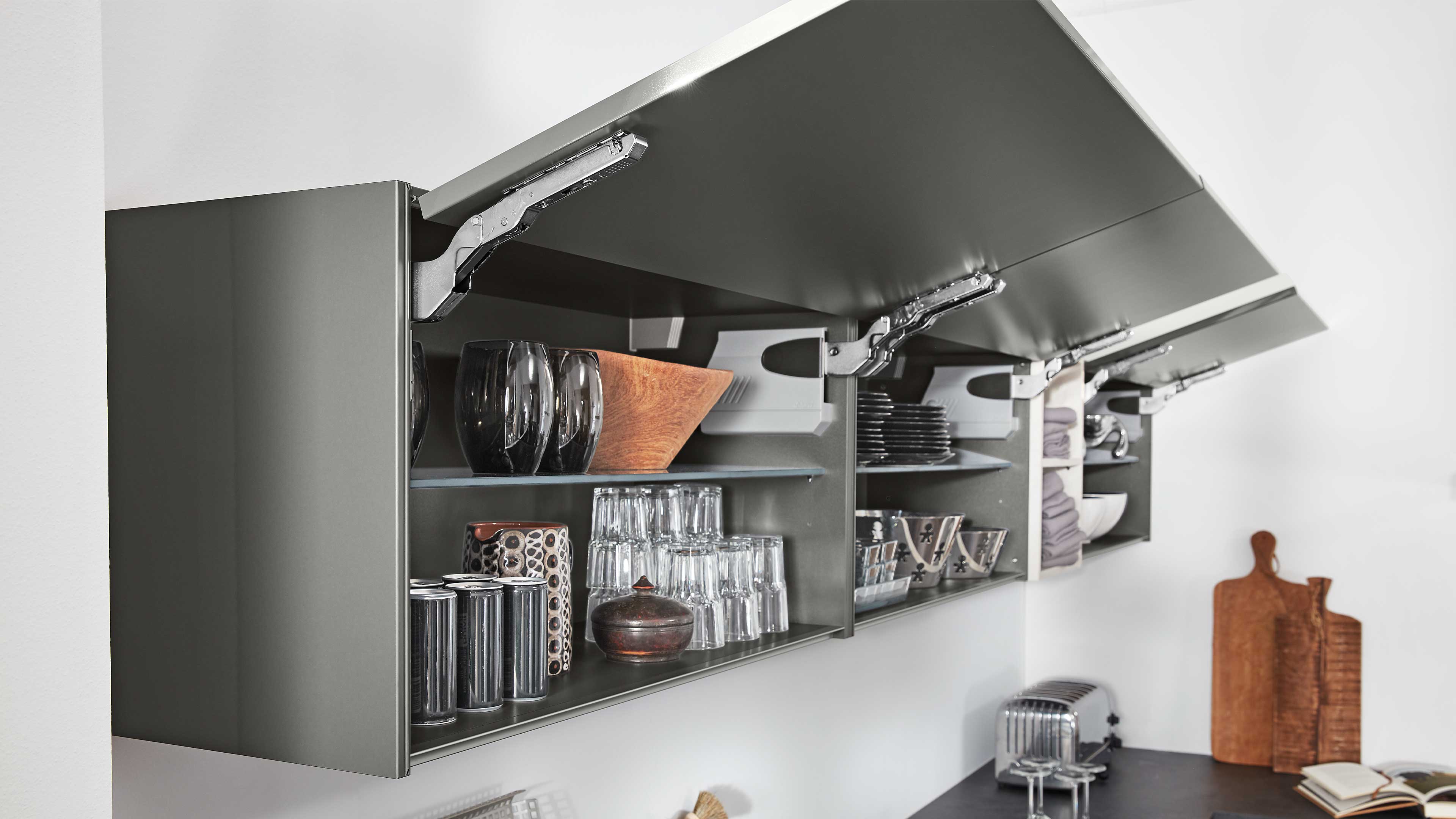 Wandhängender Küchenschrank mit aufklappbaren Fronten in mattem Grau, sichtbaren Metallbeschlägen und offenen Regalfächern mit Gläsern.