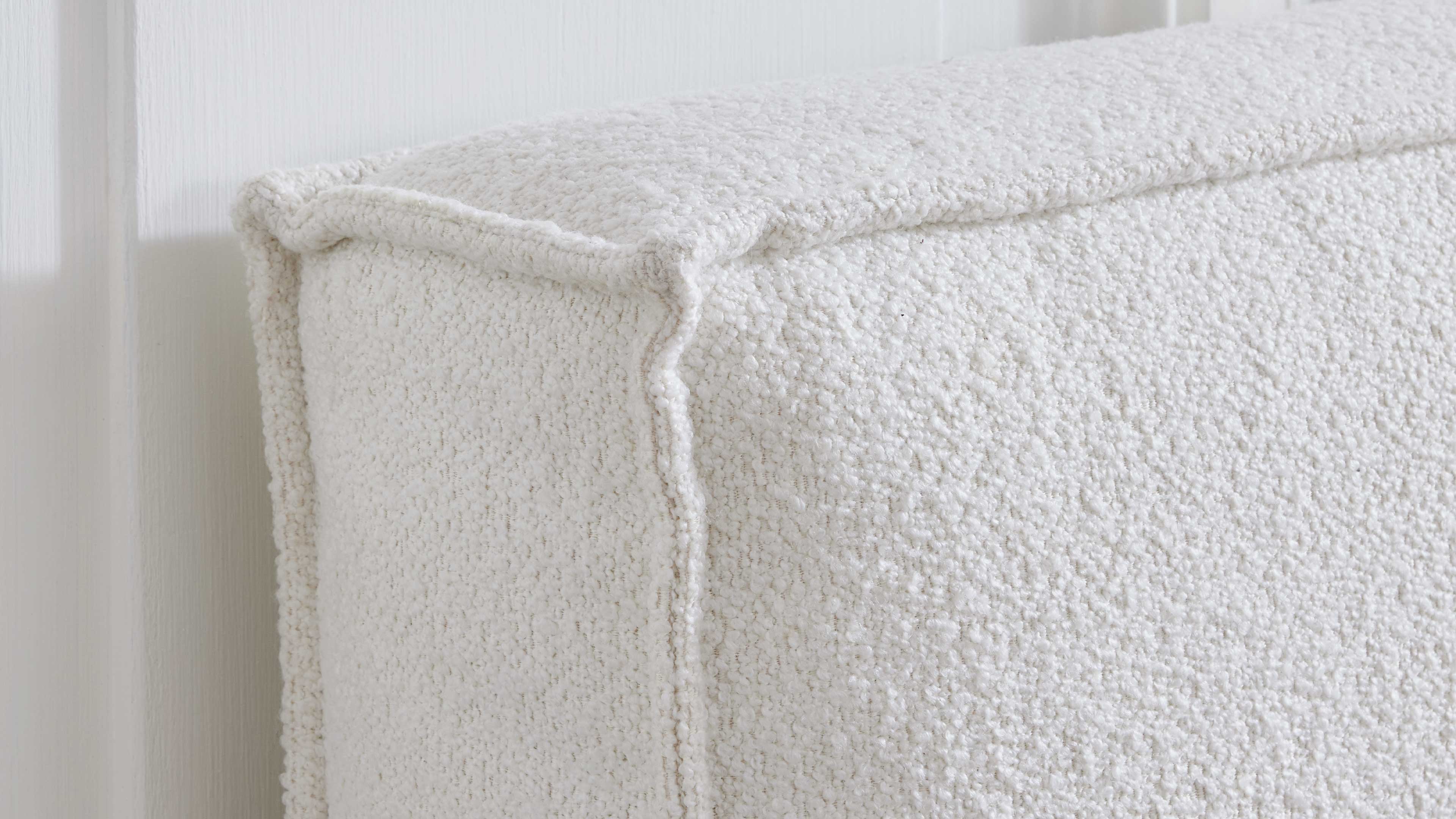 Eckige, weiße Polsterung mit grob strukturierter Bouclé-Oberfläche und betonter Nahtkante an einer Möbelecke vor heller Wand.