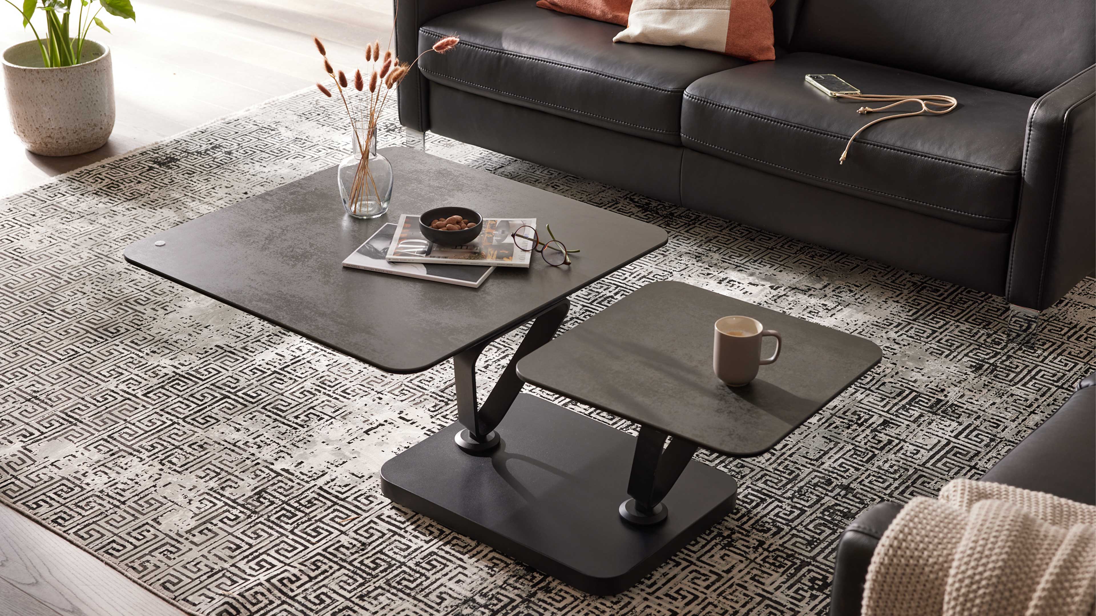 Zweistöckiger, dunkelgrauer Couchtisch mit zwei rechteckigen Platten auf schwarzer Basis, auf gemustertem Teppich vor Ledersofa.