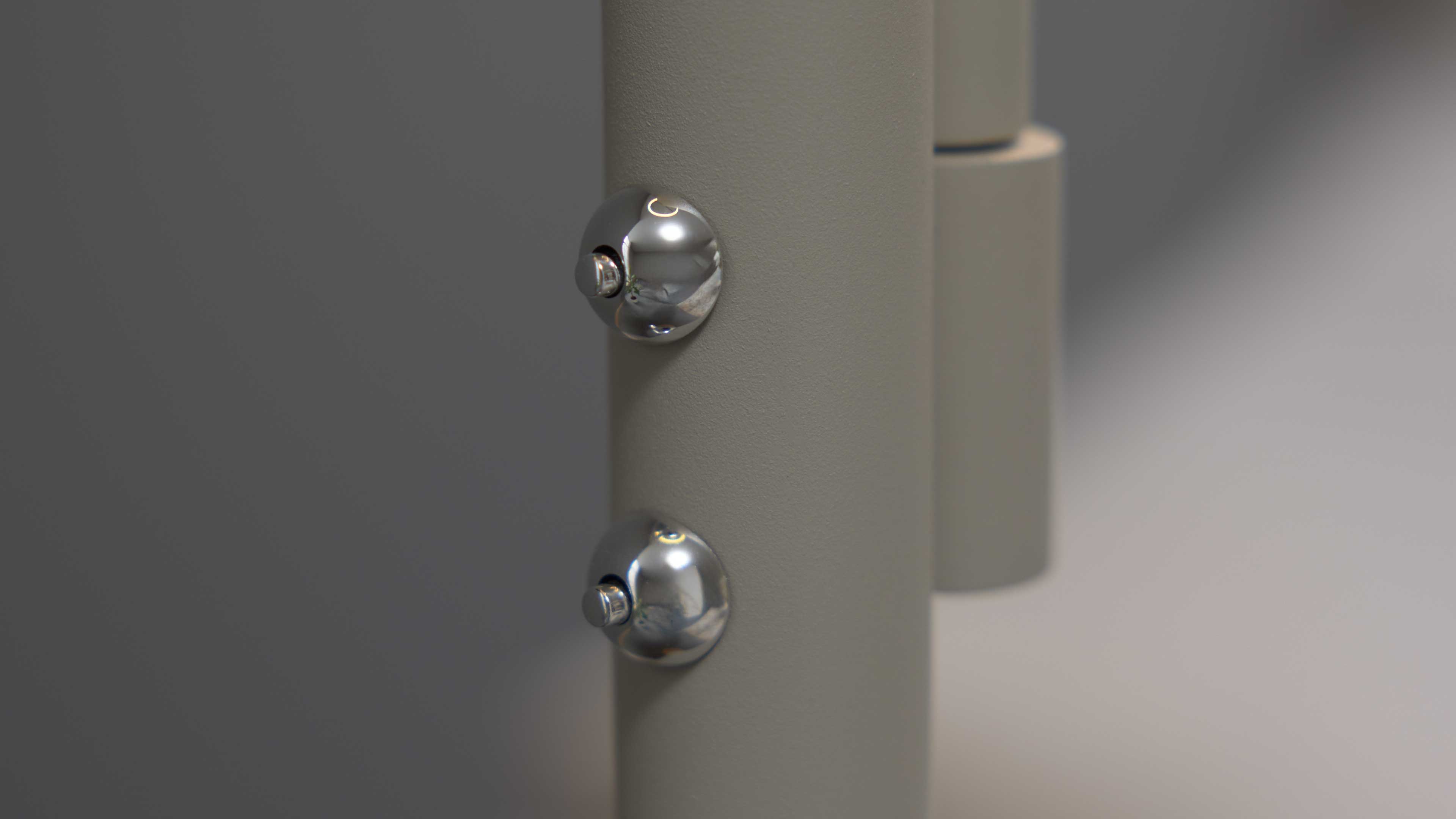 Zylindrische hellgraue Stange mit zwei runden verchromten Schraubkappen, rechts angedocktes Scharnierteil.