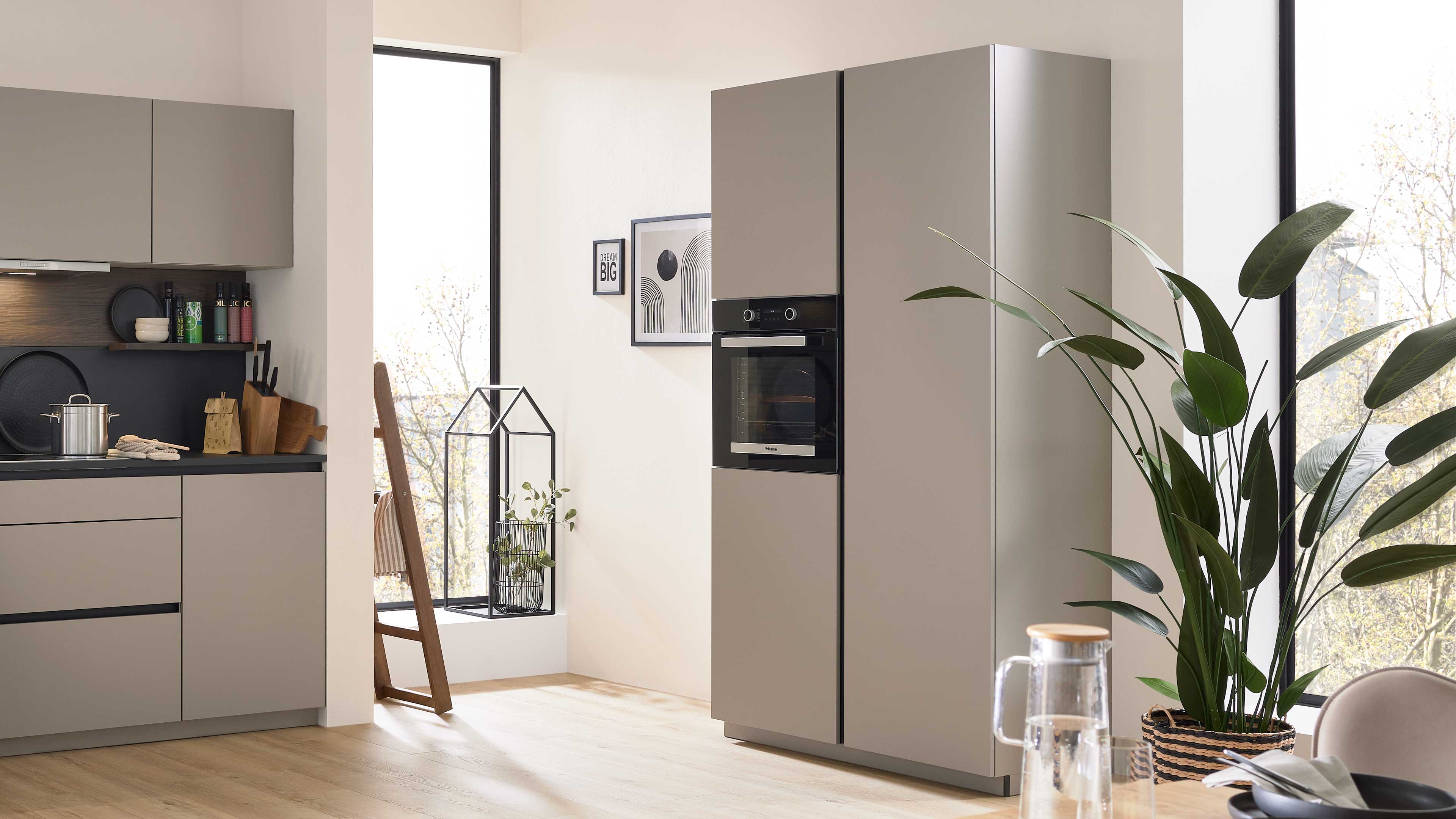 Hoher beige-grauer Küchenschrank mit integriertem Backofen in einer hellen Küche, rechts große Topfpflanze am Fenster.