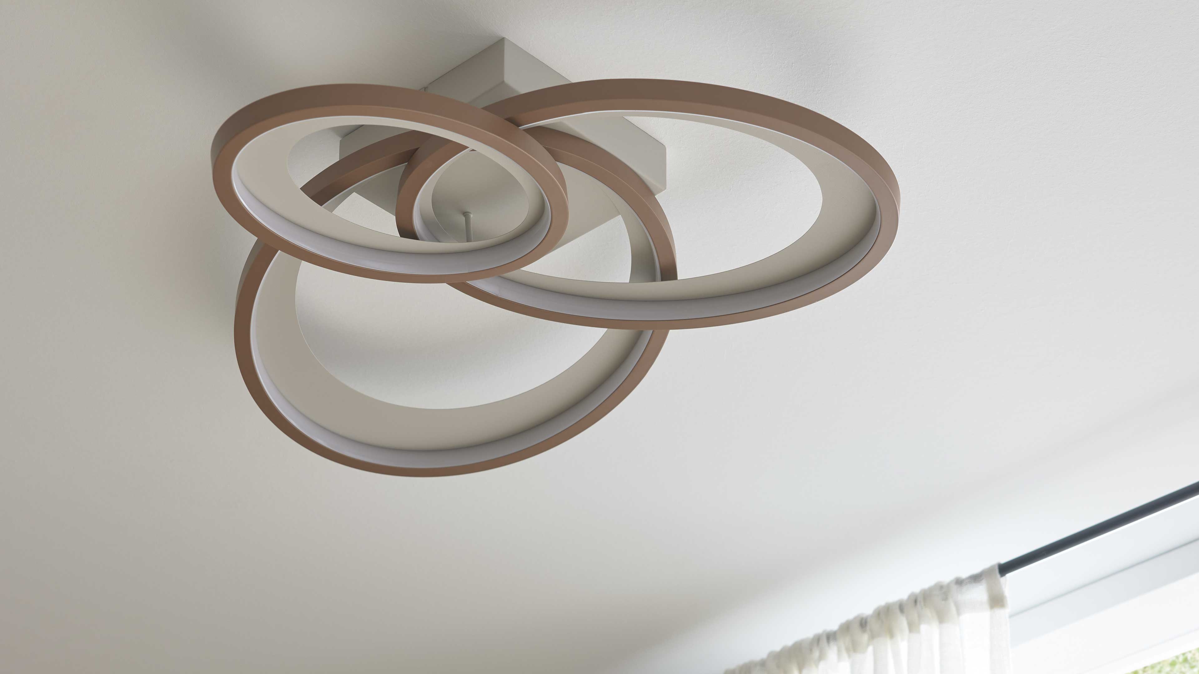 Deckenleuchte mit drei verschlungenen kreisförmigen Leuchtringen mit braunem Rand an eckiger Halterung, Vorhangstange und weißer Vorhang.