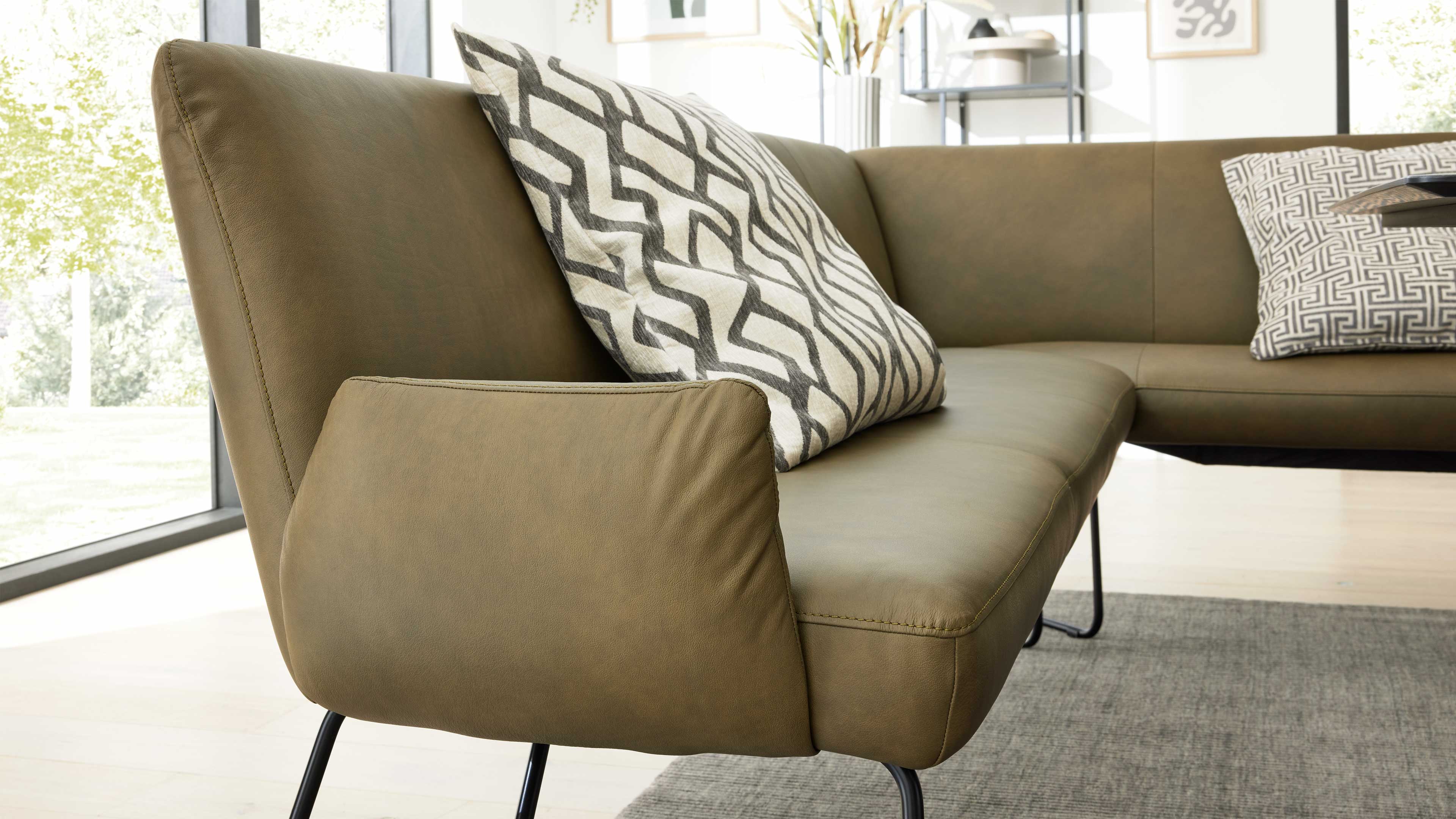 Ecksofa in grünbraunem Leder mit schwarzen Metallfüßen, zwei gemusterten Kissen und grauem Teppich in hellem Raum.