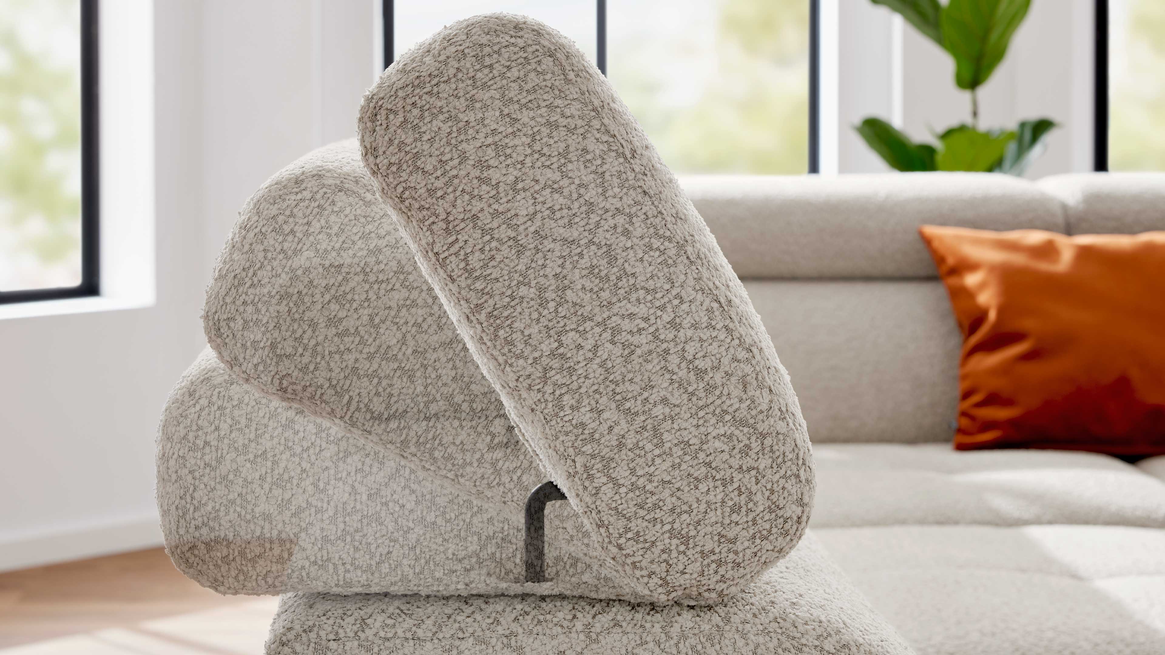 Nahaufnahme eines hellbeigen, strukturierten Sofarückens mit verstellbaren Kopfstützen in drei Positionen, orangenes Kissen im Hintergrund.