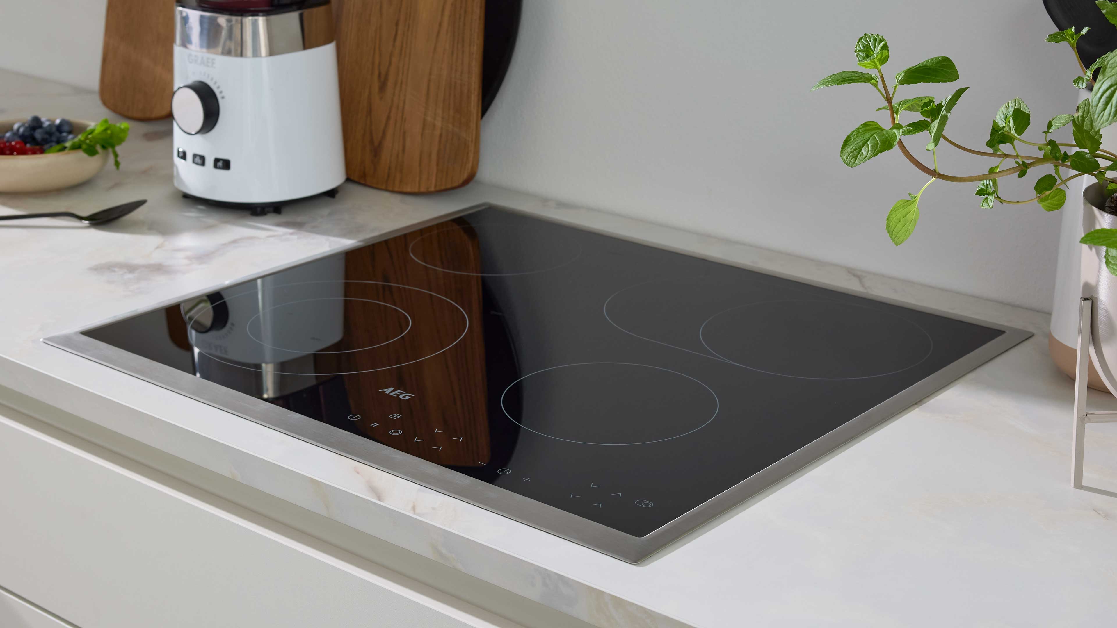 Schwarzes Glaskeramik-Kochfeld mit Edelstahlrahmen in weißem Marmor-Arbeitsplatteneinbau, links Mixer und Holzbretter, rechts Topfpflanze.