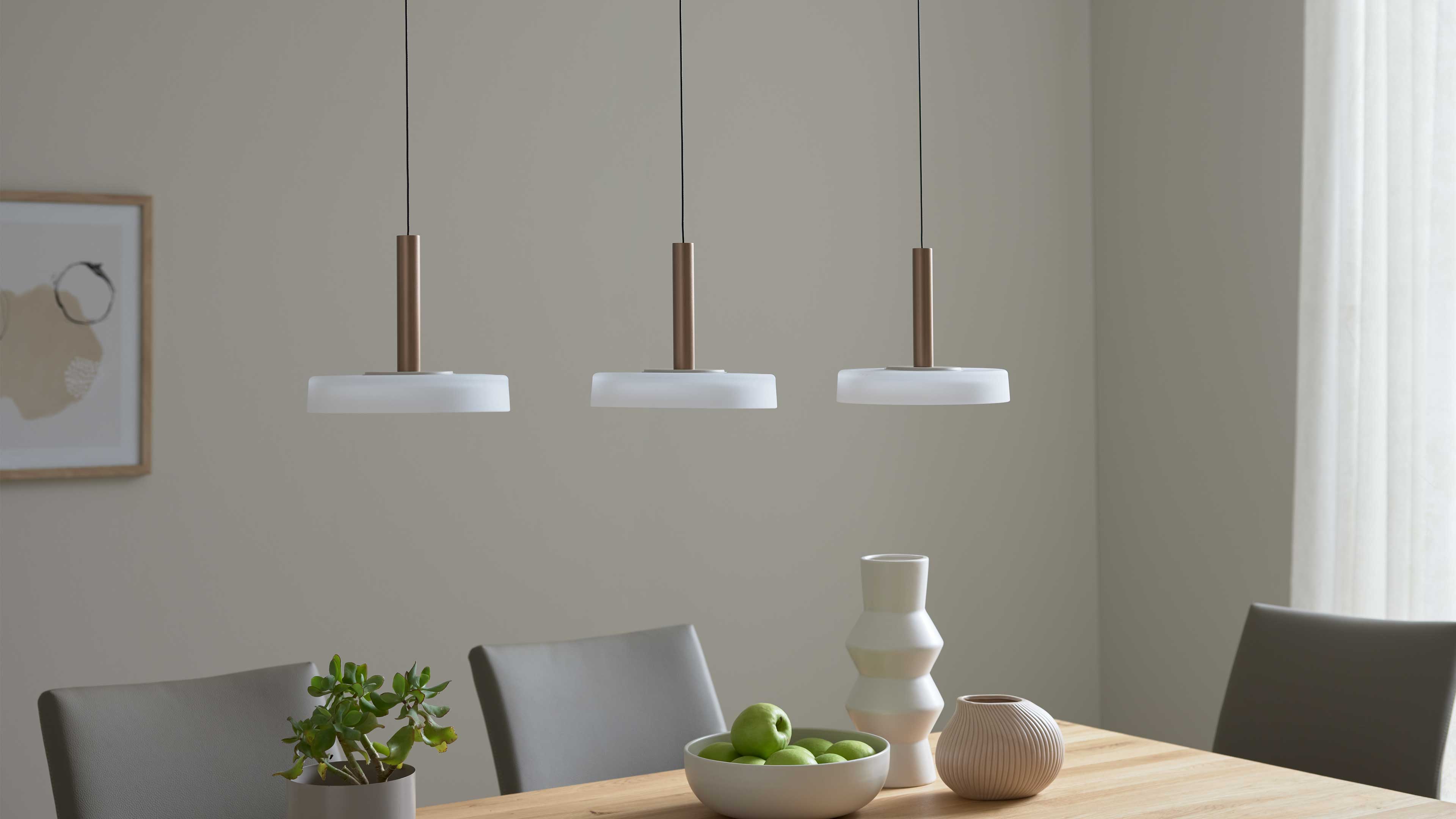 Drei hängende Pendelleuchten mit bronzefarbenem zylindrischem Stiel über einem Holztisch mit Schale grüner Äpfel, Vasen und Stühlen.