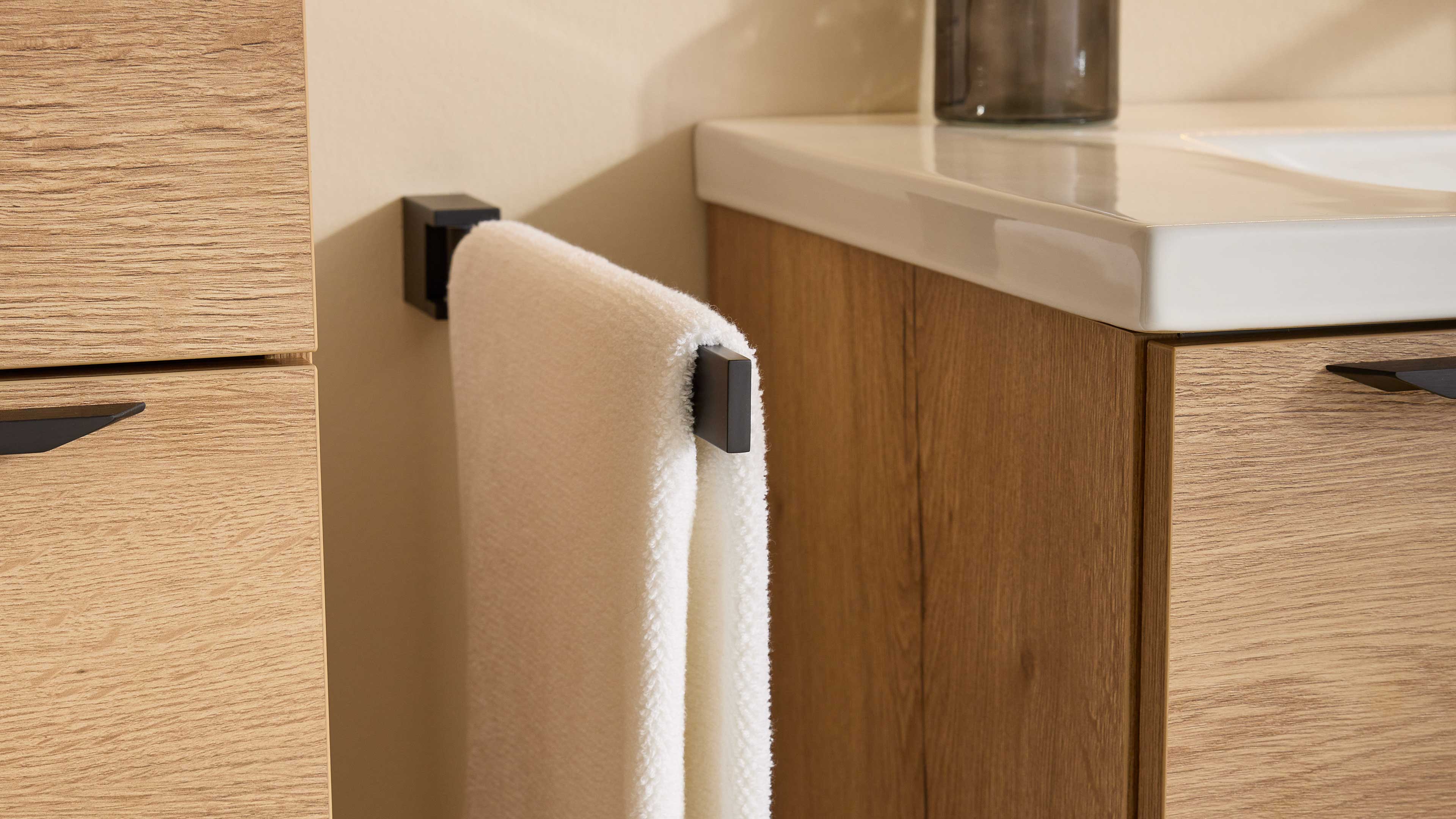 Weißes Handtuch hängt an schwarzem rechteckigem Handtuchhalter zwischen hellen Holzschränken neben weißer Waschtischplatte.
