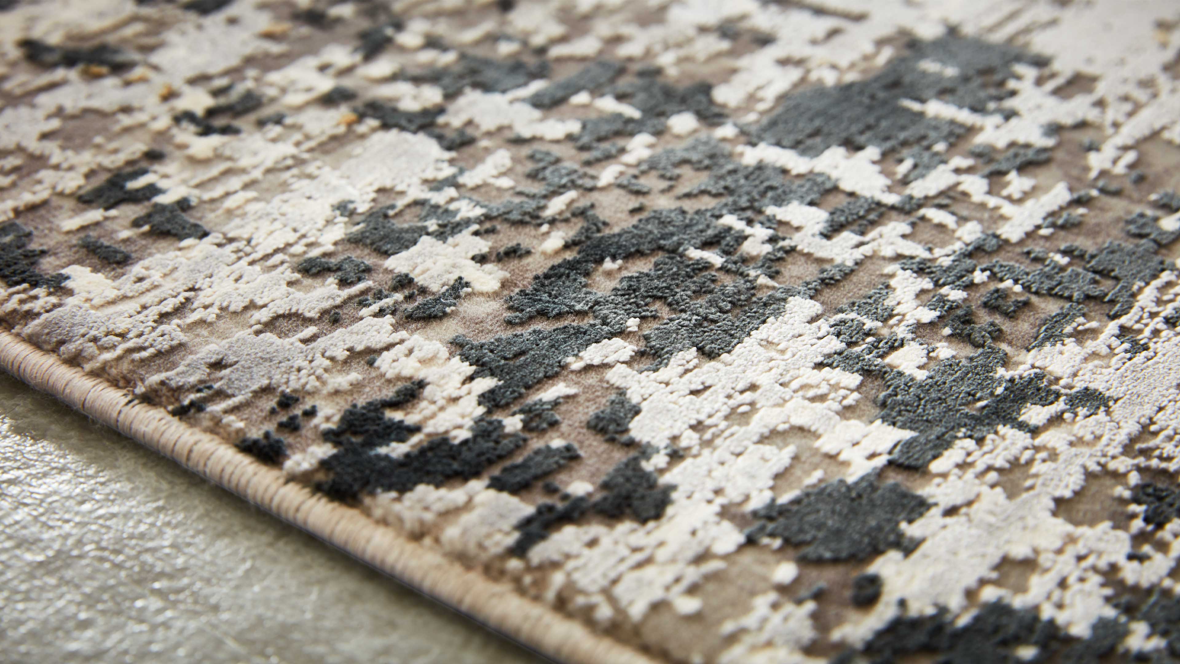 Nahaufnahme eines Teppichs mit beigen, creme- und dunkelgrauen reliefartigen Flormustern, sichtbarem eingefasstem Rand und Boden.