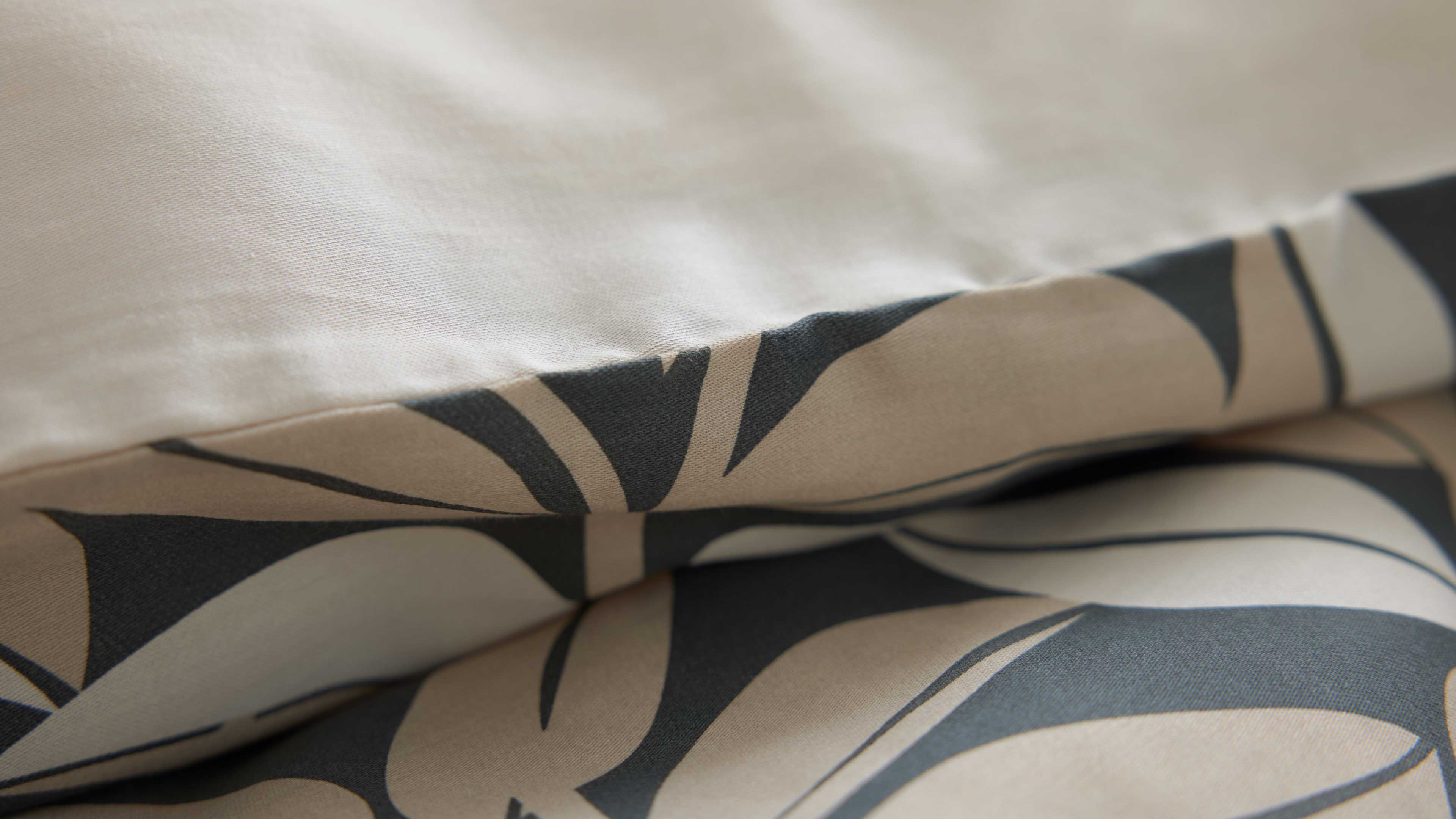 Gefalteter beiger Stoff mit großflächigem dunkelgrauem Blattmuster im Vordergrund und einer cremefarbenen Kante oben.