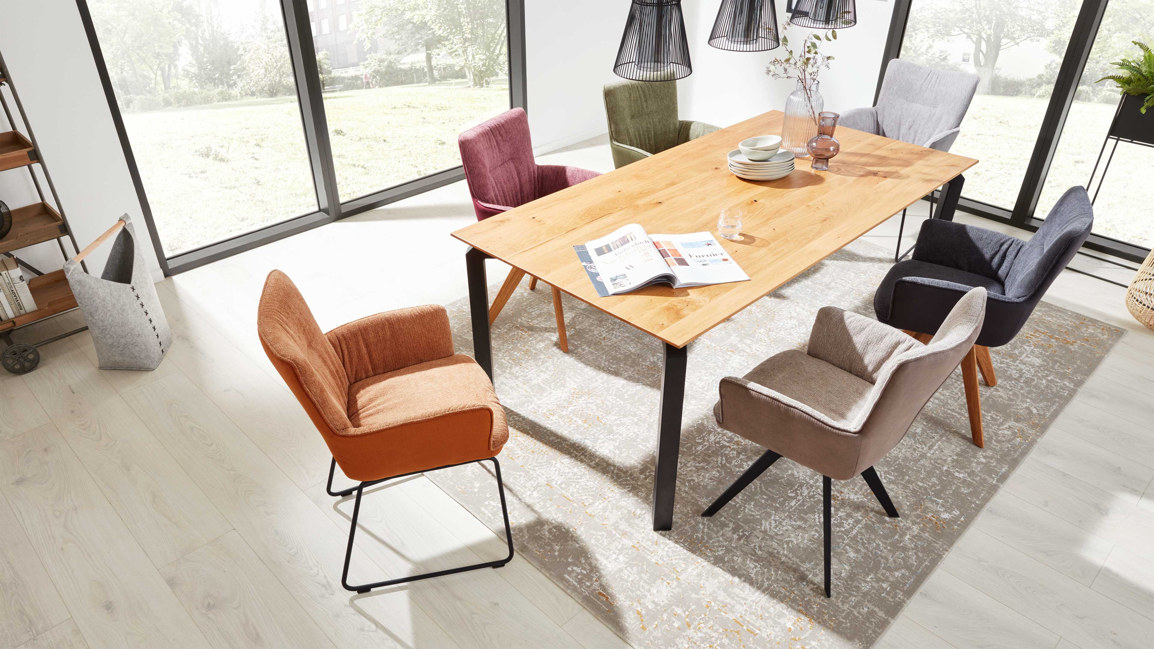 Holztisch mit schwarzen Metallbeinen, umgeben von sechs bunten Sesseln auf Teppich vor großen Panoramafenstern.