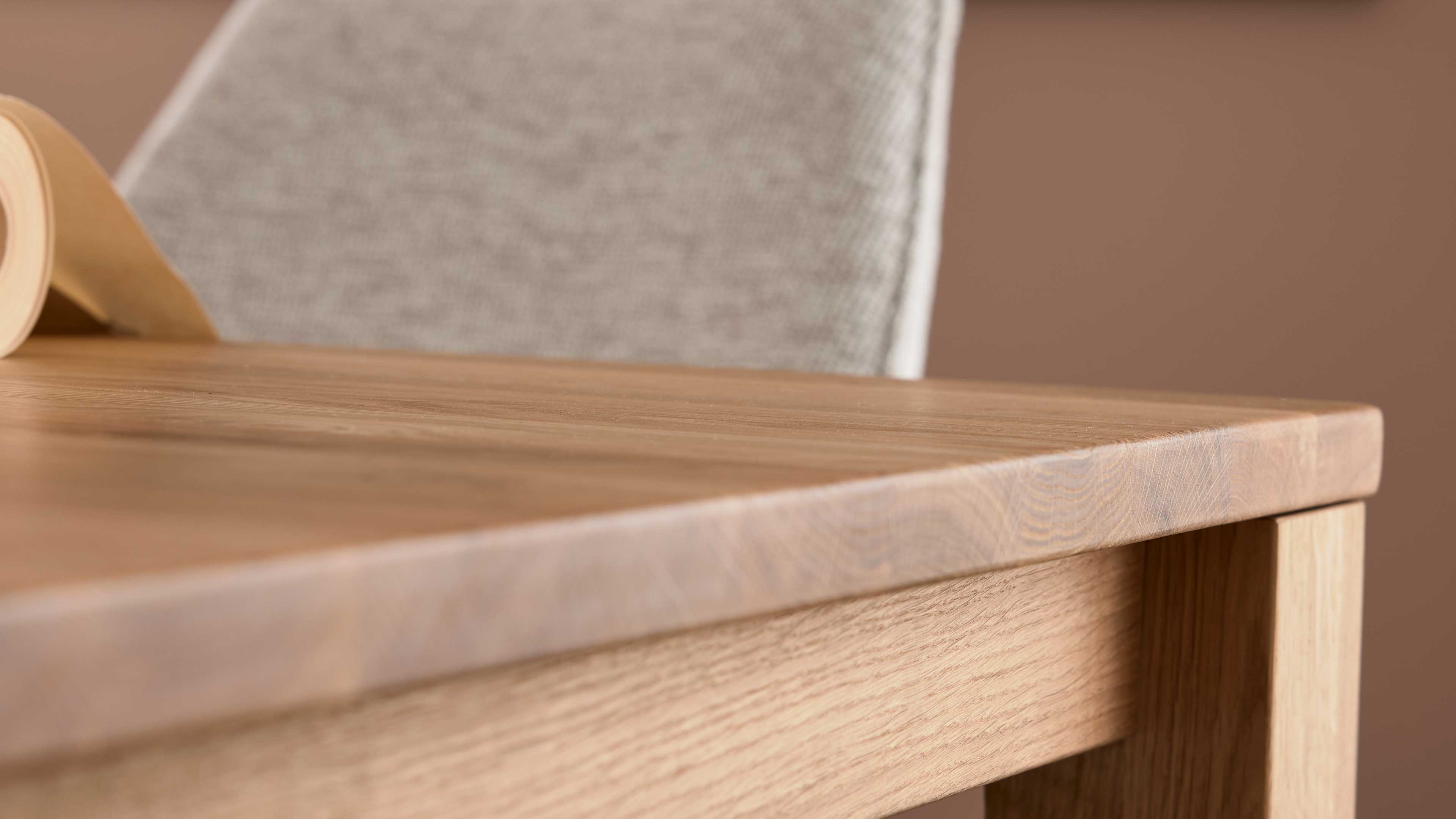 Holz-Tischplatte mit sichtbarer Maserung und abgeschrägter Kante, im Hintergrund ein grauer Stoffstuhlrücken und aufgerolltes Papier.