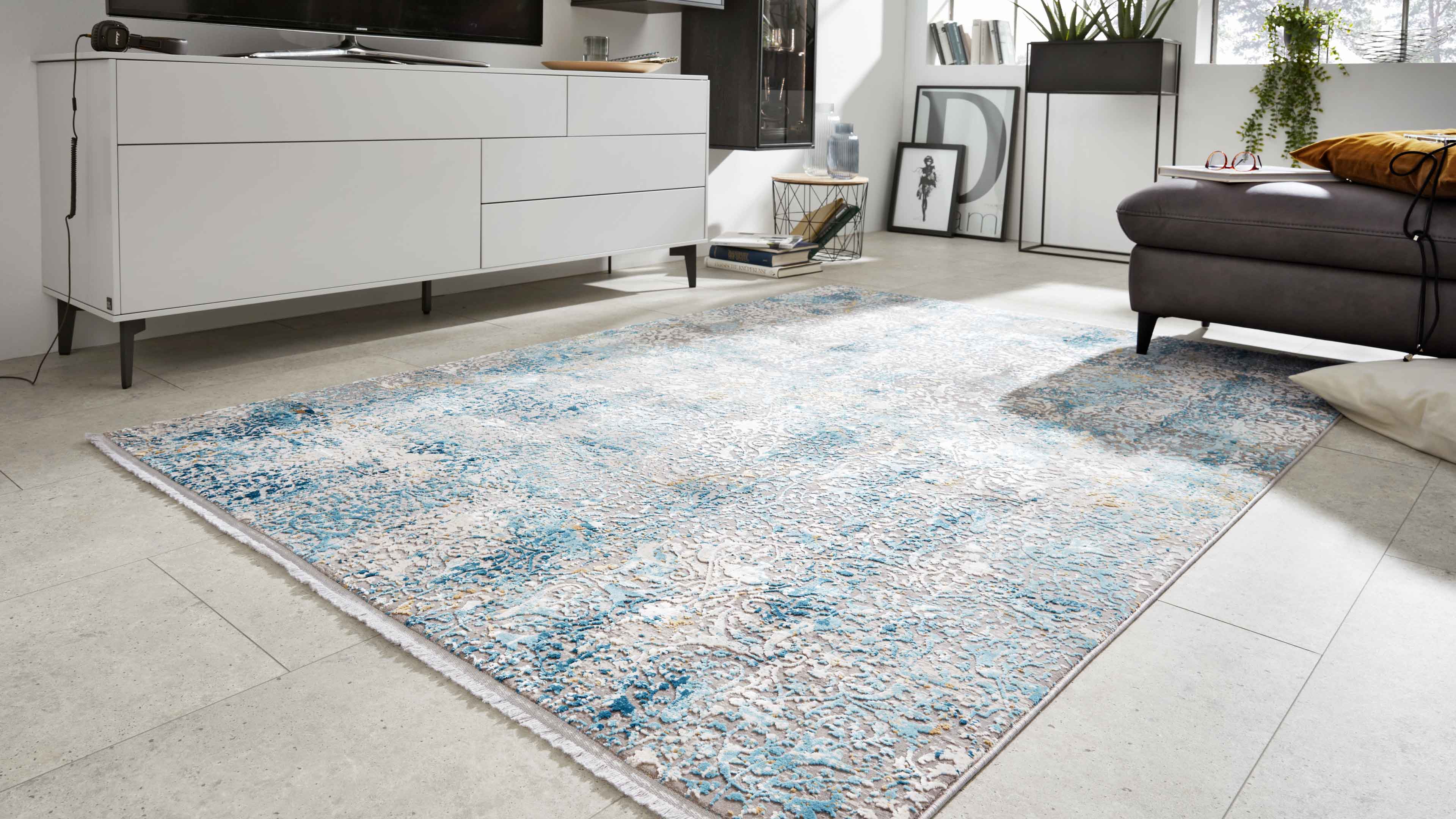 Großflächiger Teppich mit abstraktem blau-beige Muster auf hellen Bodenfliesen, daneben weiße Kommode und graues Sofa.