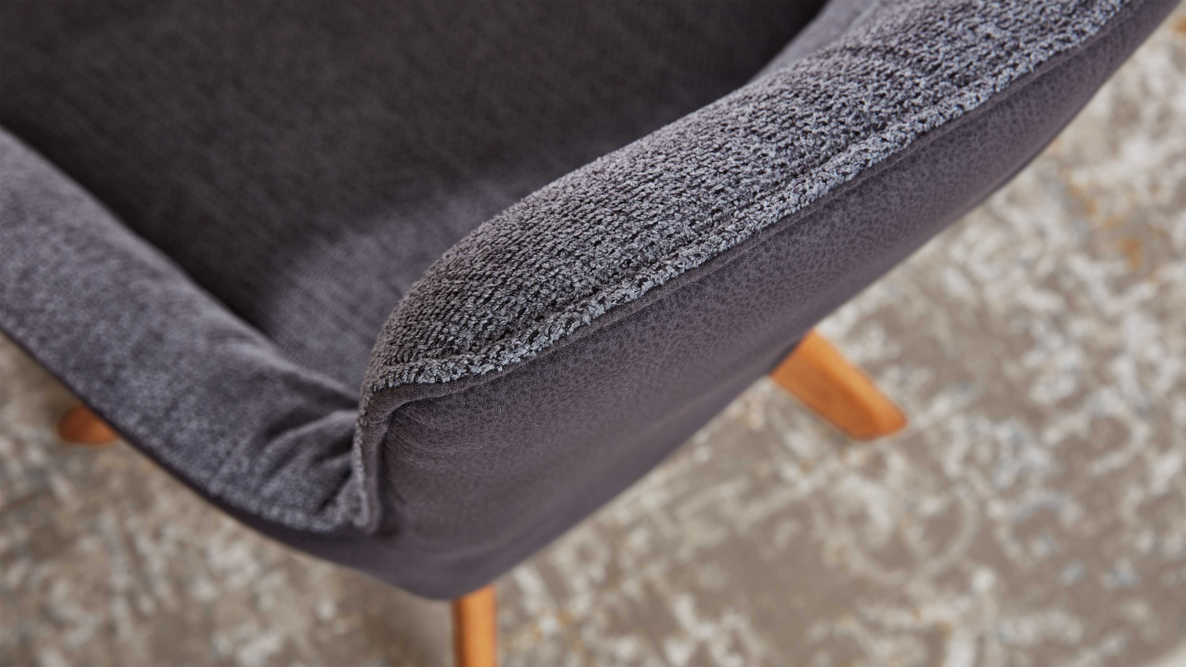 Grauer, gepolsterter Sesselarm mit strukturierter Stoffkante, sichtbares helles Holzbein auf gemustertem Teppich.