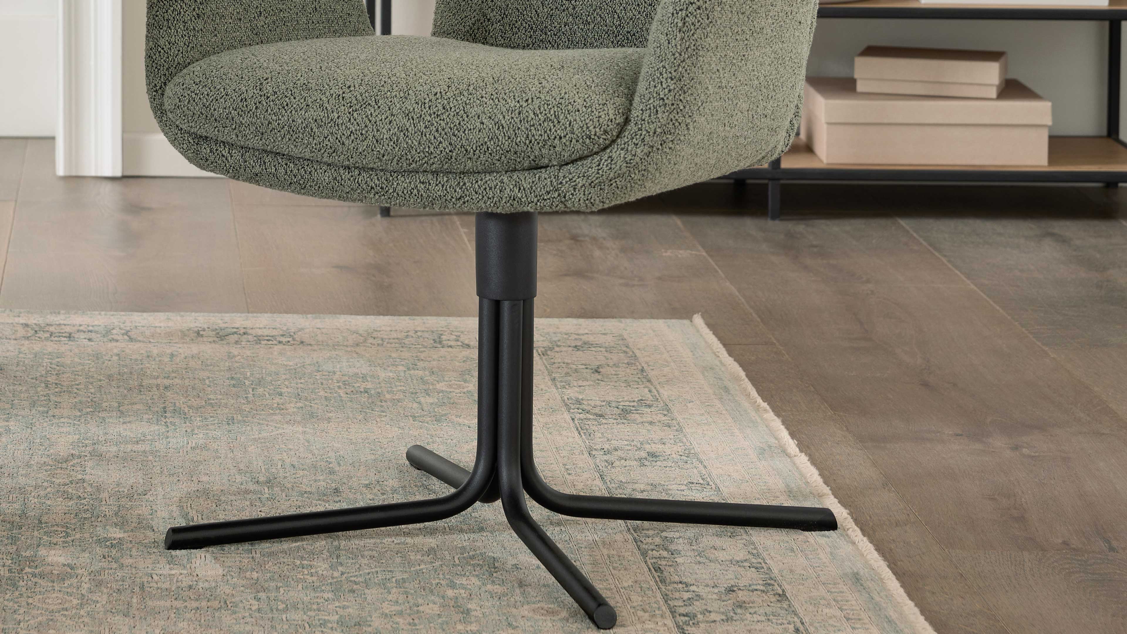 Grüner, bezogener Sessel mit rundem Sitz und schwarzem vierarmigem Metallfuß auf Teppich und Holzboden, Regal mit Kartons im Hintergrund.