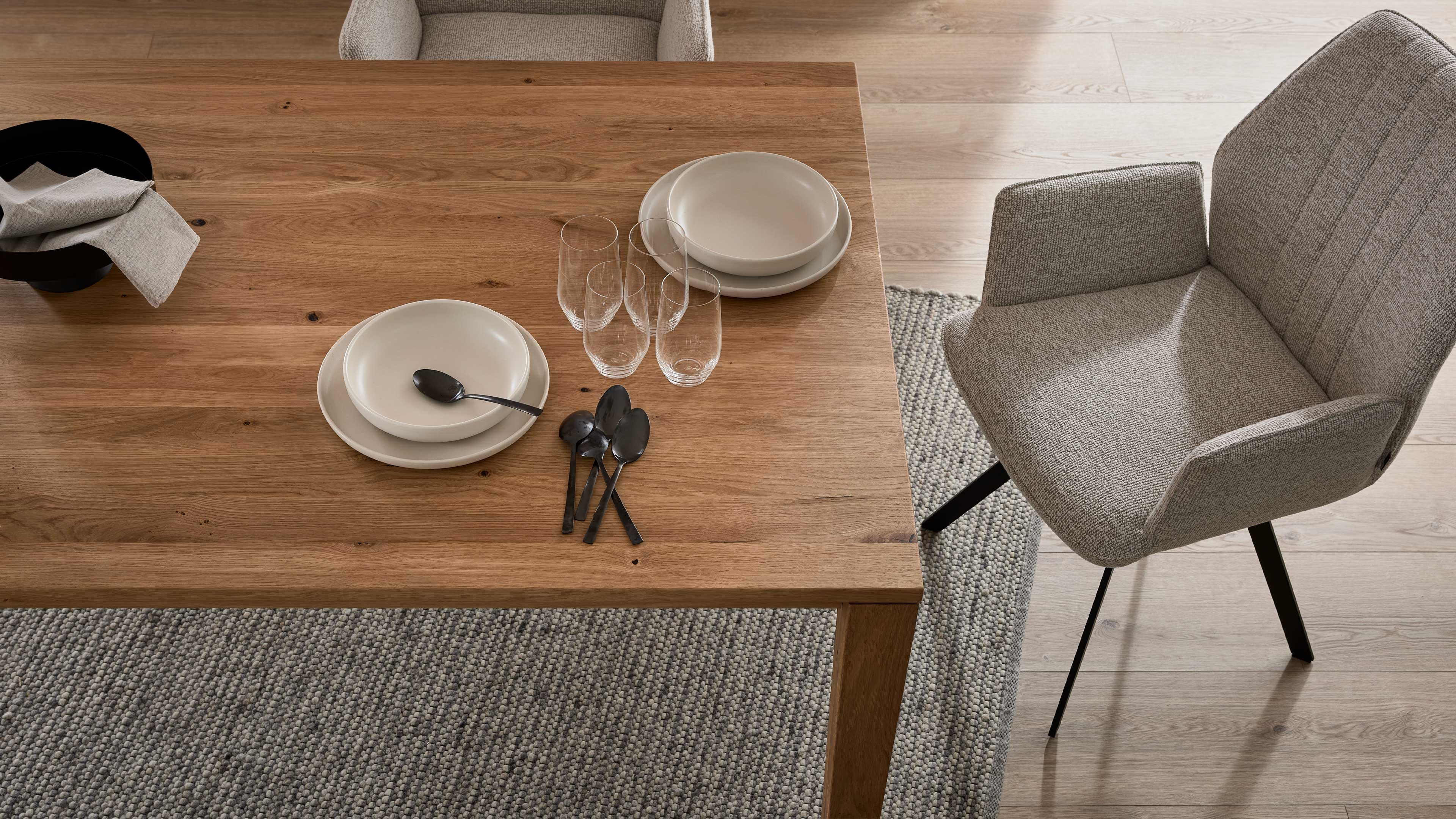 Holztisch mit gestapelten weißen Tellern, vier Gläsern und schwarzem Besteck, rechts ein grau bezogener Armlehnstuhl auf Teppich.