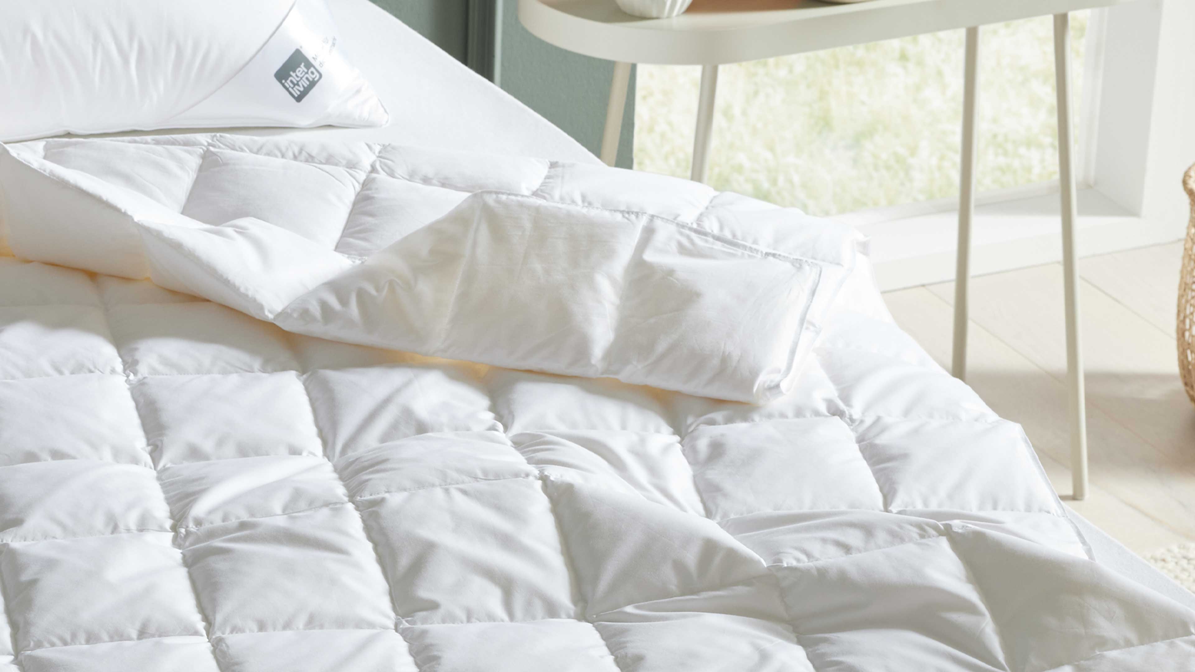 Weiß gesteppte Bettdecke mit sichtbarer Falte und darauf liegendem weißen Kopfkissen, im Hintergrund Tischbeine und Fenster.