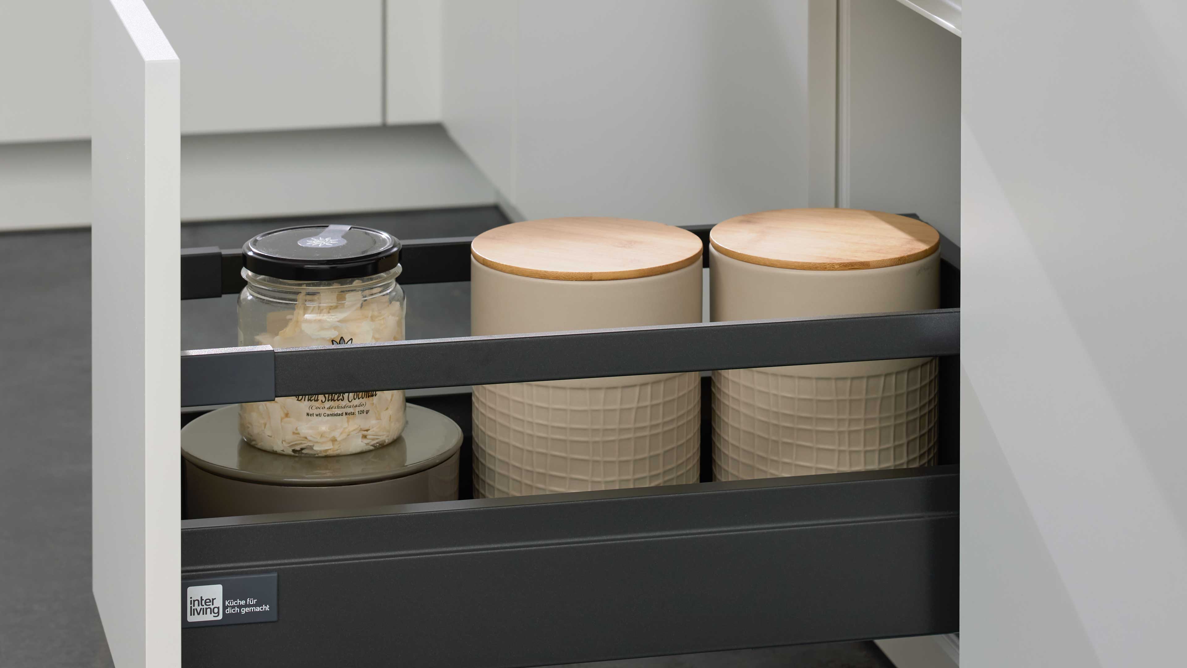 Offener grauer Schubkasten mit drei beige Vorratsdosen mit Holzdeckeln rechts und einem Glas mit schwarzem Deckel links.