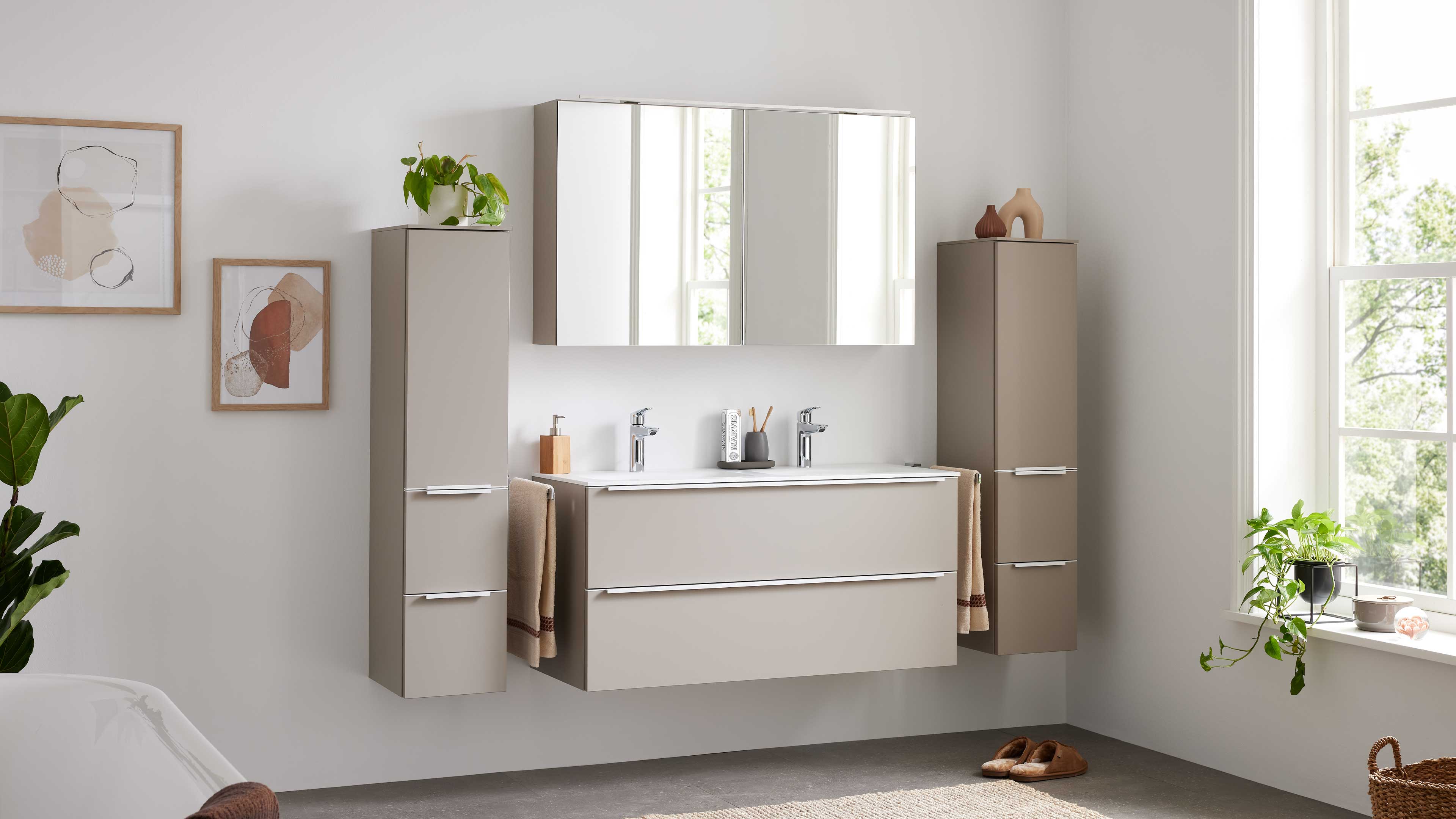 Wandhängende beige Badezimmerschränke mit Doppelwaschtisch, Spiegelschrank, zwei hohen Seitenschränken und Handtüchern.