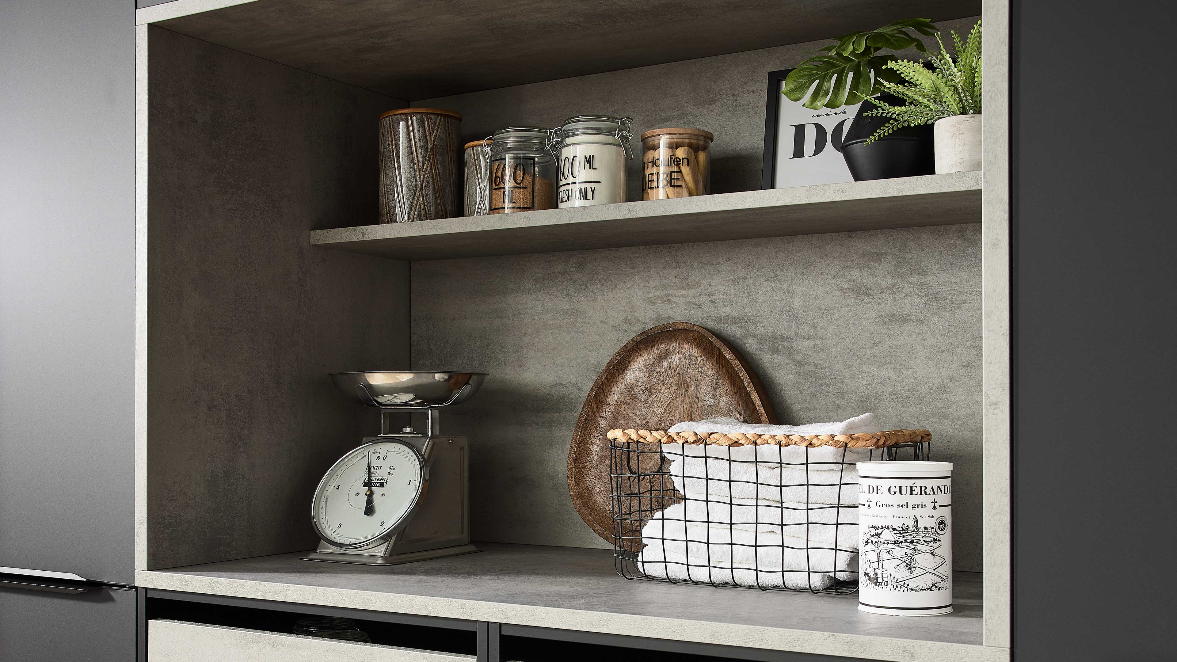 Offenes Küchenregal mit Drahtkorb gefüllt mit weißen Handtüchern, Tablett dahinter, linke Waage, obere Ablage mit Gläsern und Pflanzen.