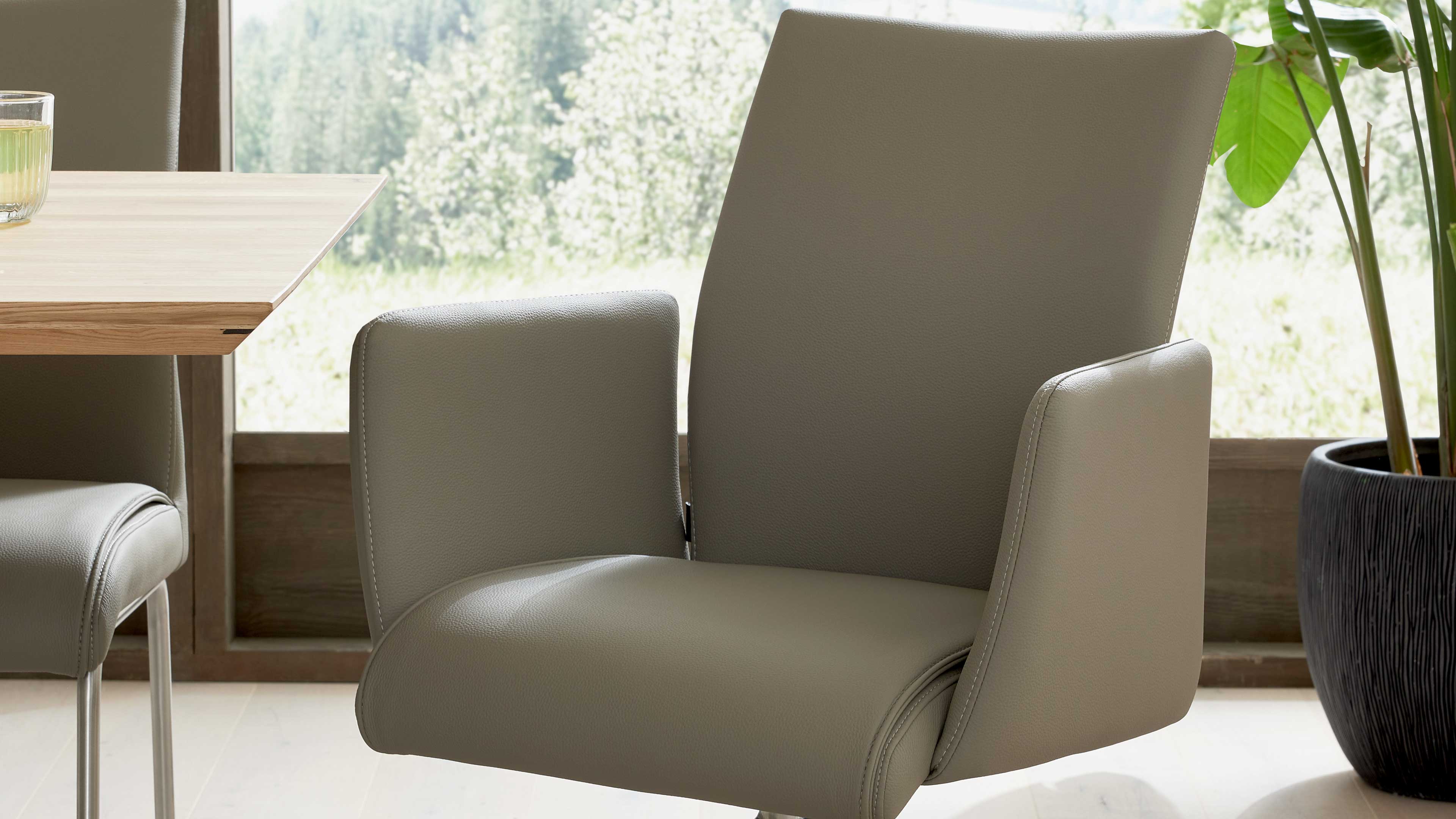 Gepolsterter graubrauner Armlehnstuhl mit breiter Sitzfläche, sichtbare Armlehnen, links Holztisch und Trinkglas, rechts Topfpflanze.