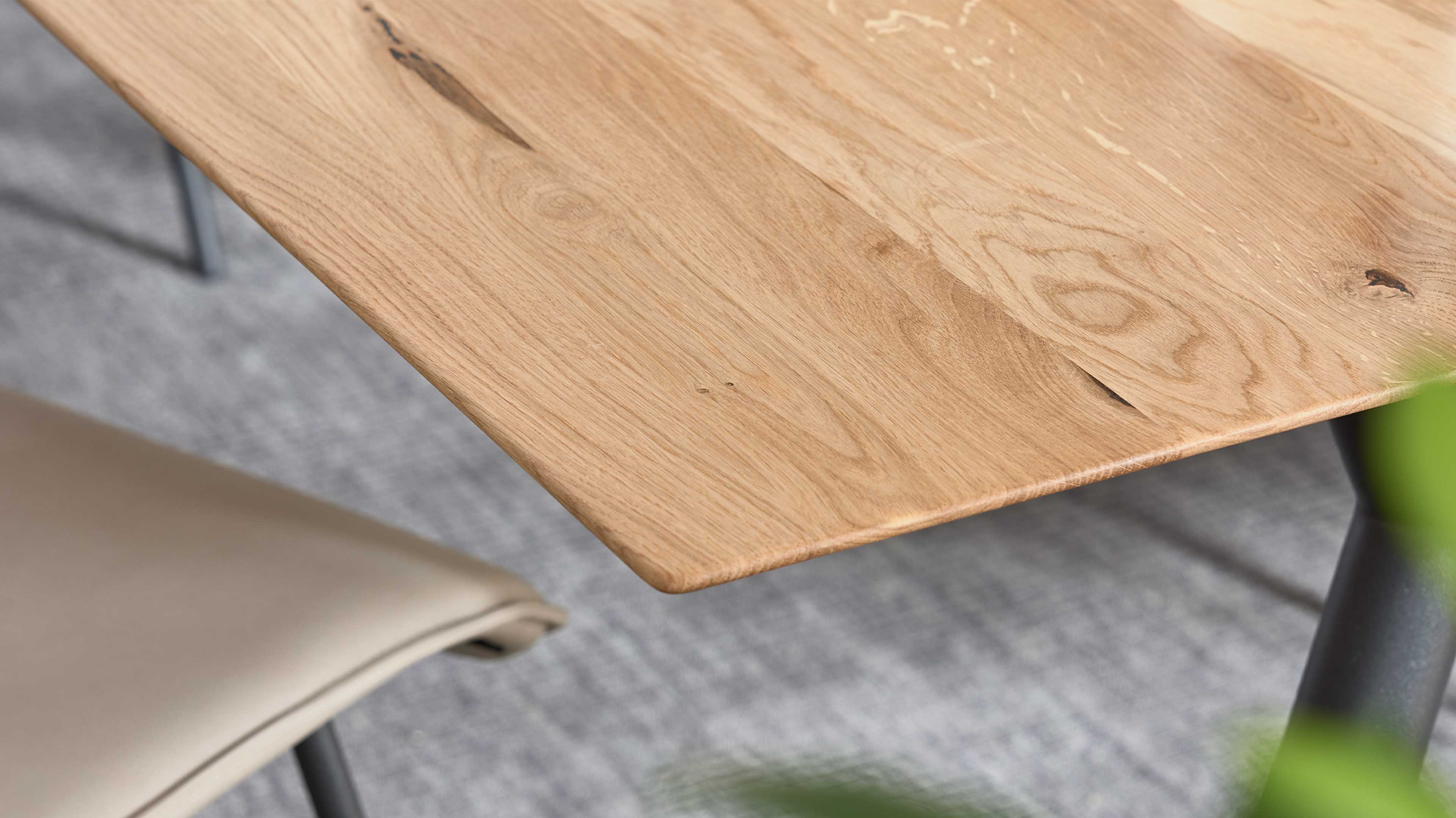 Eckige Tischplatte aus hellem Holz mit sichtbarer Maserung und Astlöchern, schwarz lackiertes Tischbein, beige Stuhl.