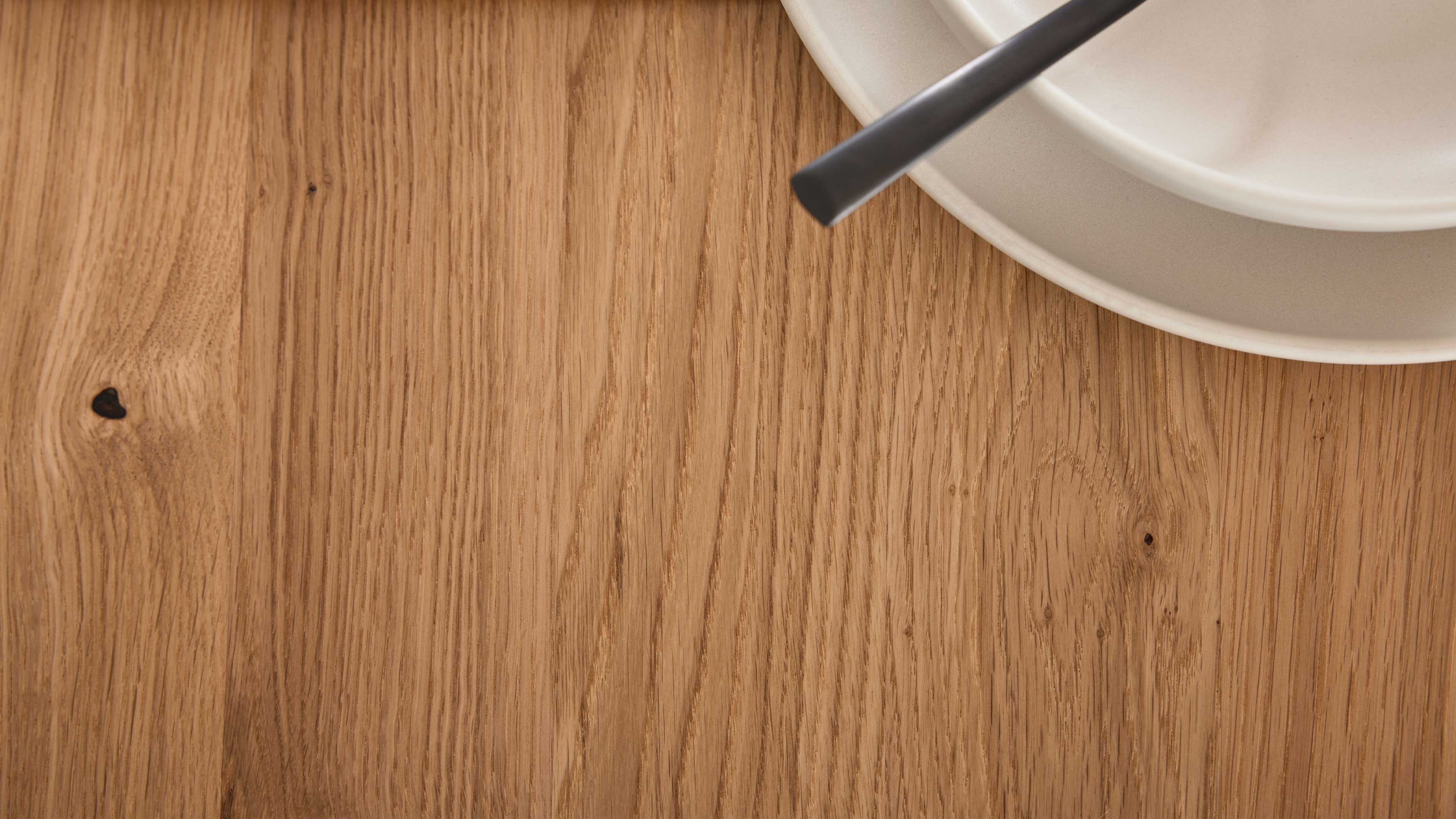 Großflächige hellbraune Eichen-Tischplatte mit sichtbarer Maserung, oben rechts zwei beige Teller und ein schwarzer Besteckgriff.