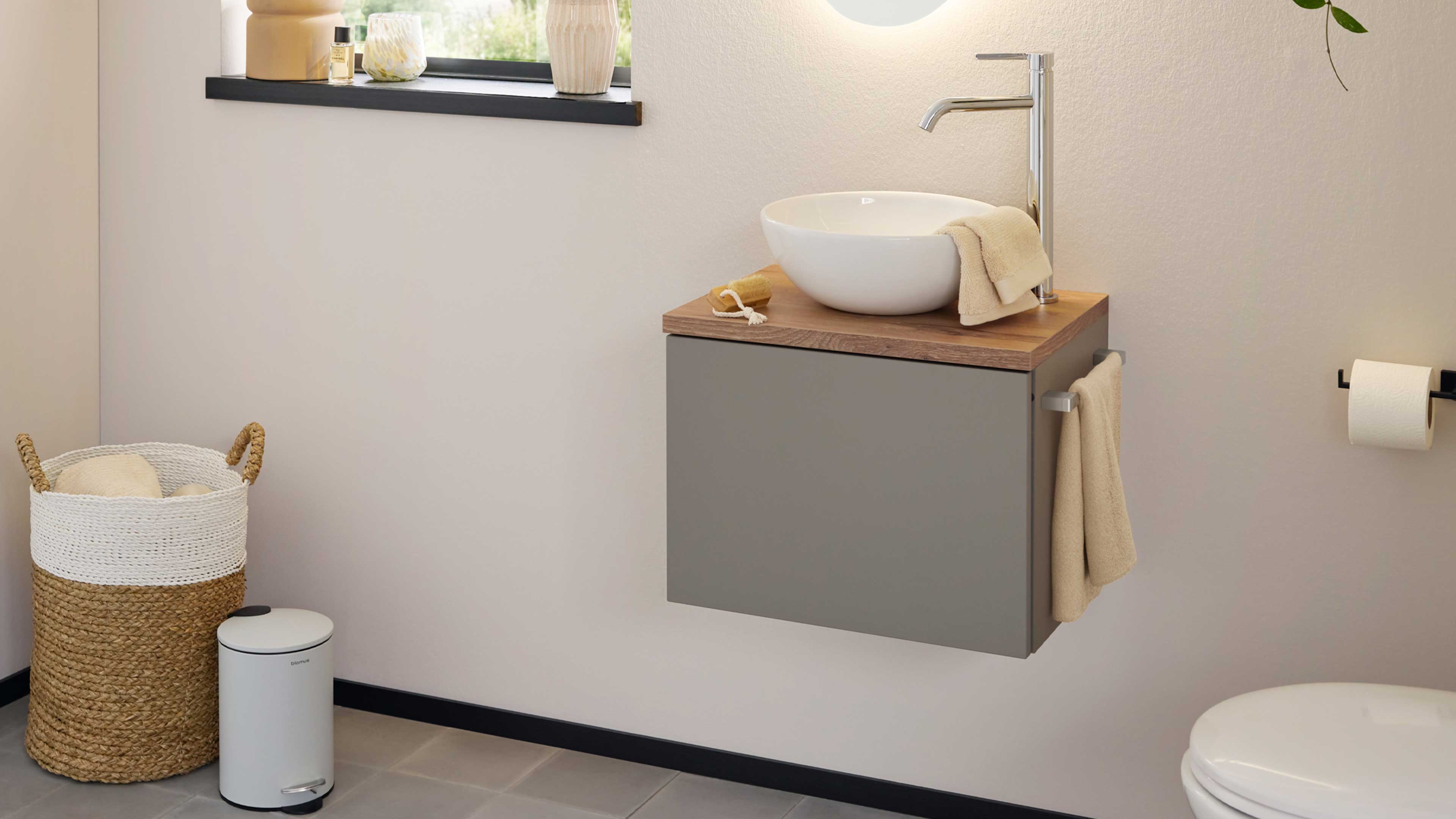 Wandhängender grauer Badezimmerschrank mit Holzplatte, weißer Aufsatzwaschschale, Chromarmatur und beigefaltetem Handtuch.