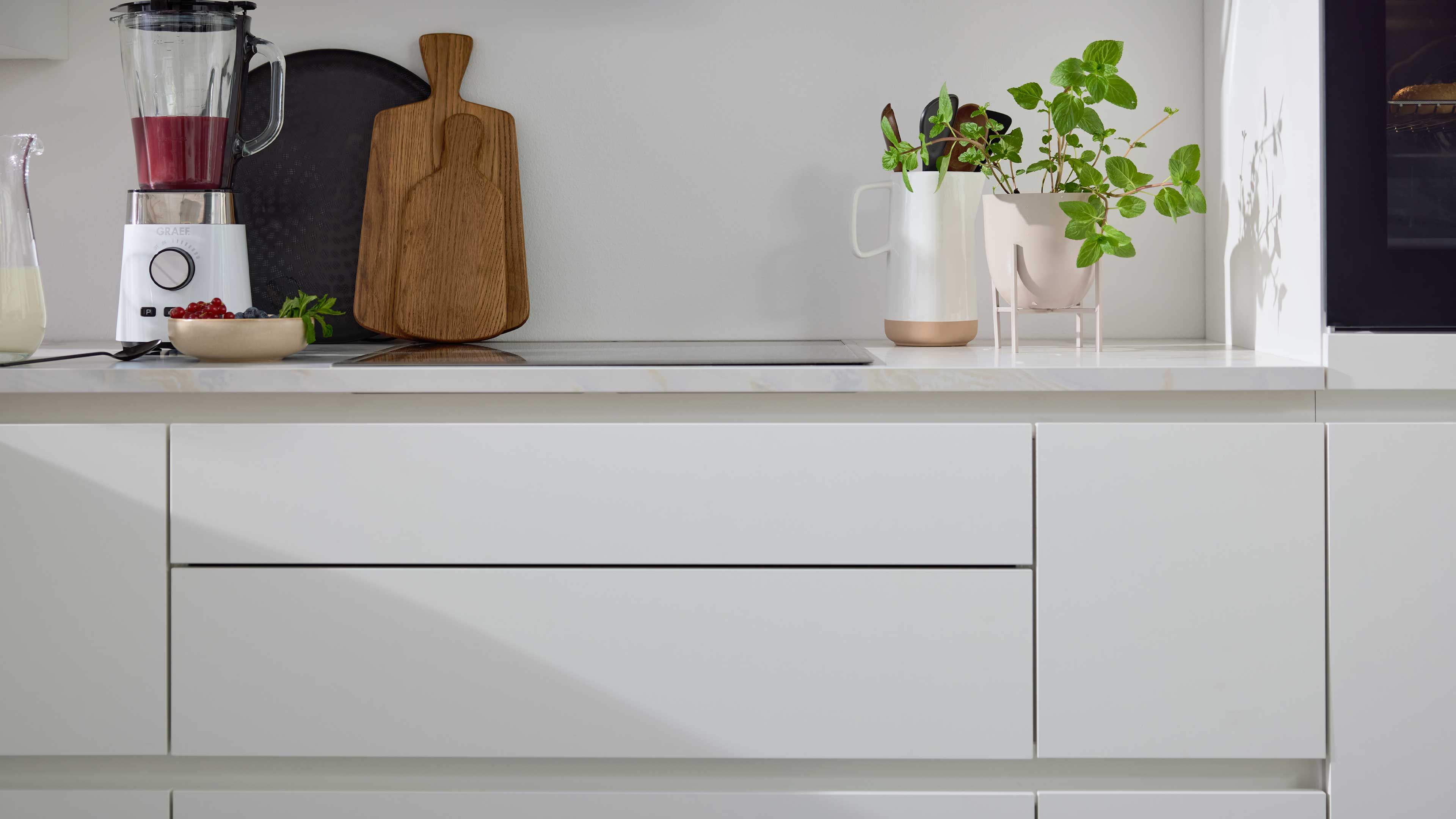 Weiße Küchenfronten mit Arbeitsplatte, links Mixer, Holz-Schneidebretter und Schale mit Beeren, rechts Kanne und Topf mit grüner Pflanze.