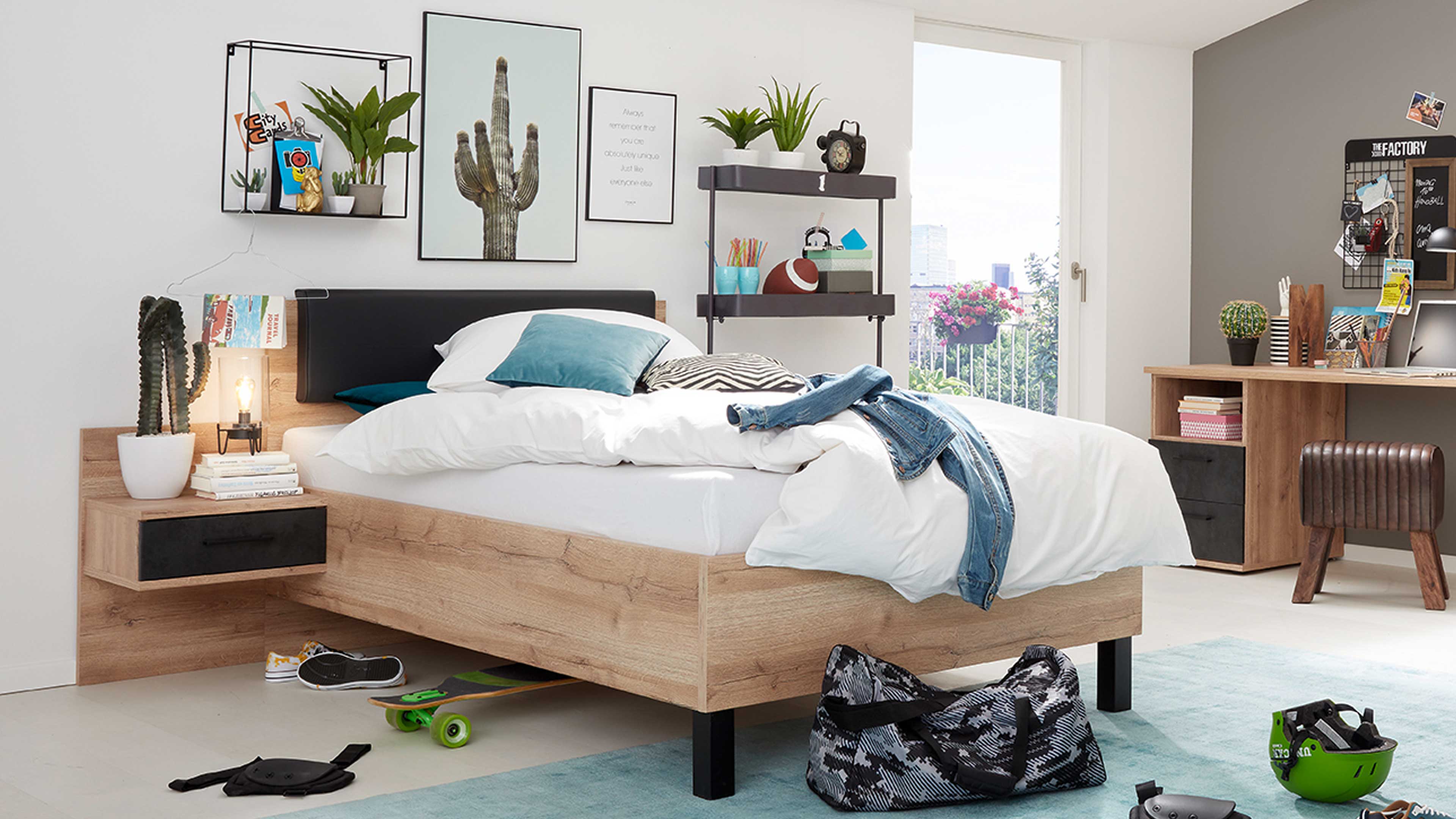 Bett aus hellem Holz mit schwarzem Kopfteil, weißer Bettwäsche und Jeansjacke, links Nachtkonsole mit Kaktus und Lampe.