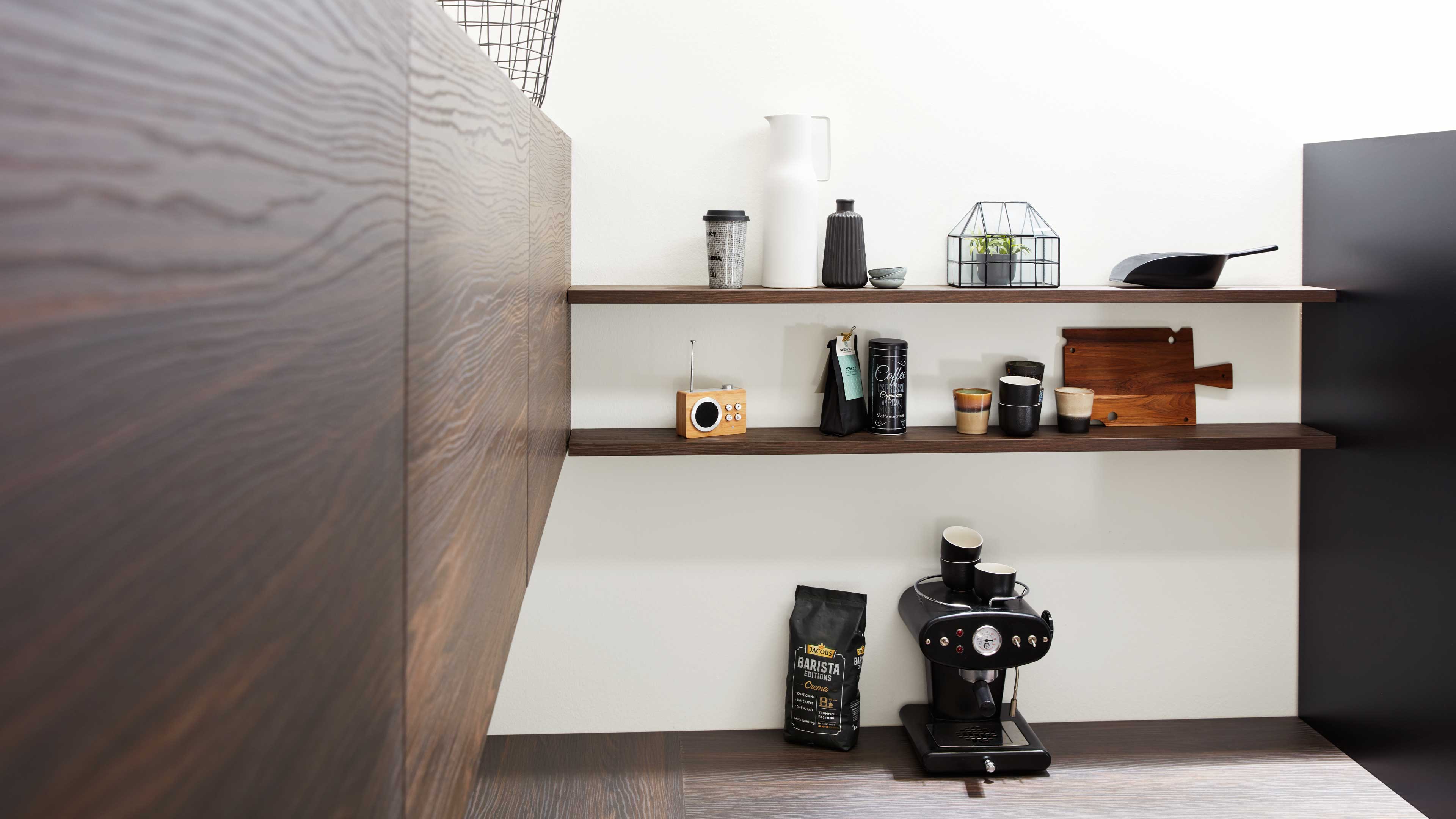 Offene dunkle Holzregale über Arbeitsfläche mit schwarzer Espressomaschine, Kaffeepaket, Tassen, Holzbrettern und kleinem Pflanzenglas.